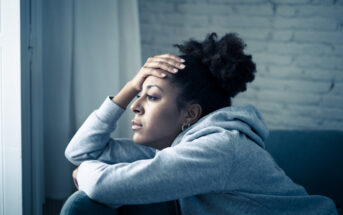 A person wearing a gray hoodie sits on a couch, resting their head on their hand while gazing out a window with a thoughtful expression. The room has a brick wall and curtains.