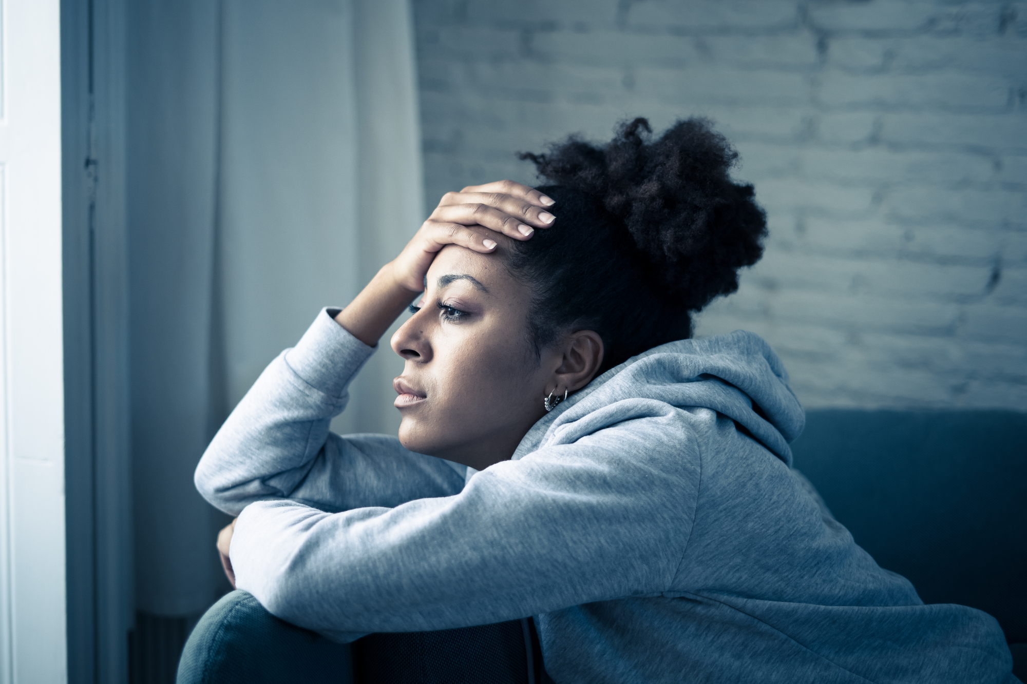 A person wearing a gray hoodie sits on a couch, resting their head on their hand while gazing out a window with a thoughtful expression. The room has a brick wall and curtains.