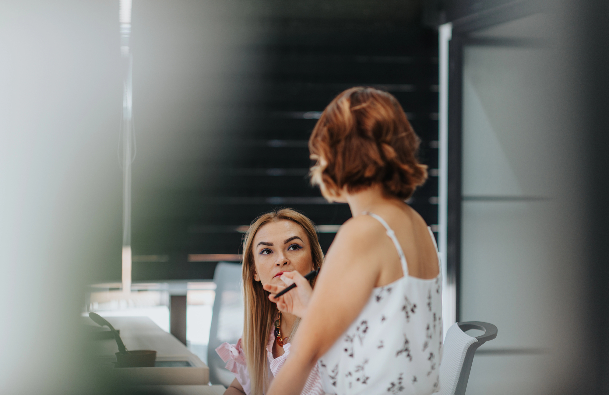 Two women are engaged in conversation at a desk in a modern office setting. The woman sitting is listening intently, while the woman standing gestures as she speaks. The background features a black paneled wall.