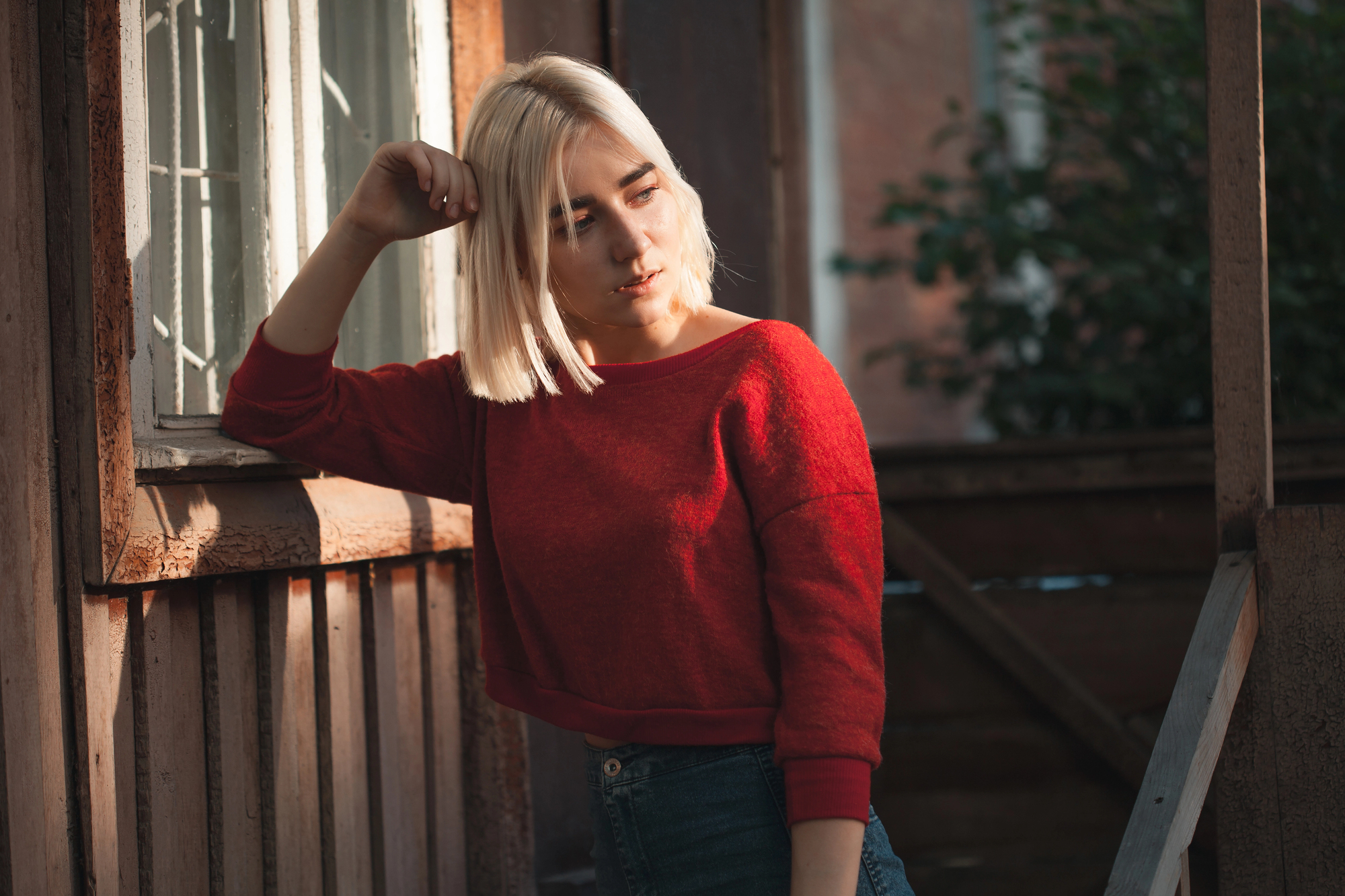 A person with blonde hair leans against a wooden building, wearing a red sweater and jeans. They pose with one hand touching their hair, bathed in sunlight, and cast a contemplative look downward.