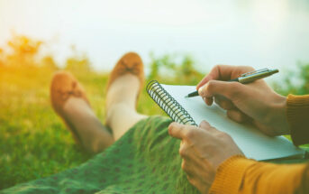 A person sits on grass, holding a pen and writing in a spiral notebook. Their legs are stretched out, wearing brown shoes. The scene is calm and sunlit, suggesting a peaceful outdoor setting.