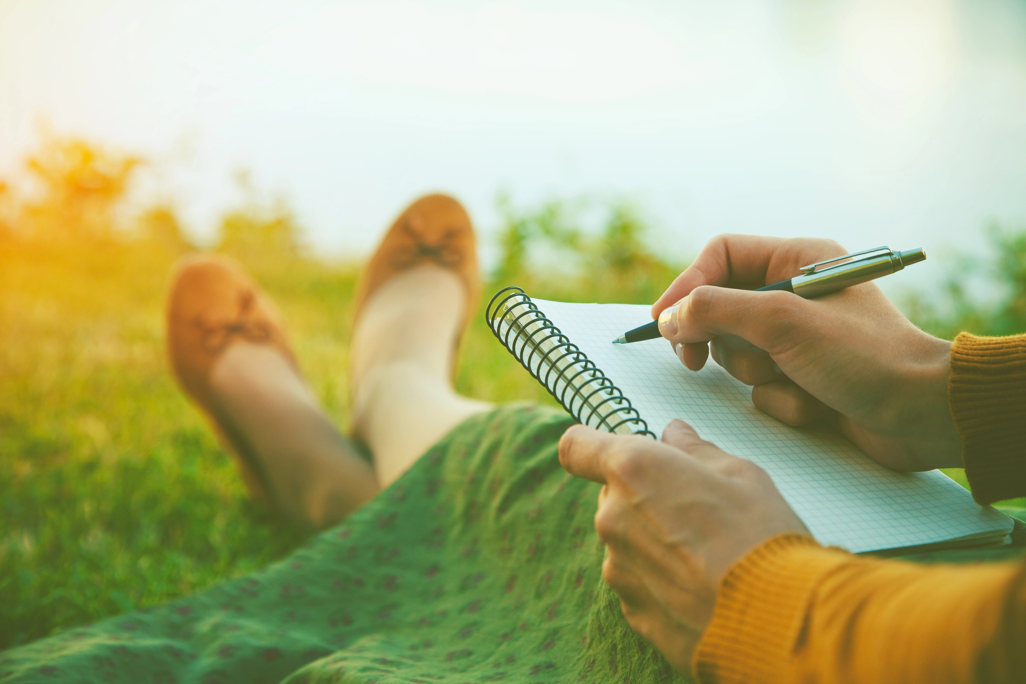 A person sits on grass, holding a pen and writing in a spiral notebook. Their legs are stretched out, wearing brown shoes. The scene is calm and sunlit, suggesting a peaceful outdoor setting.