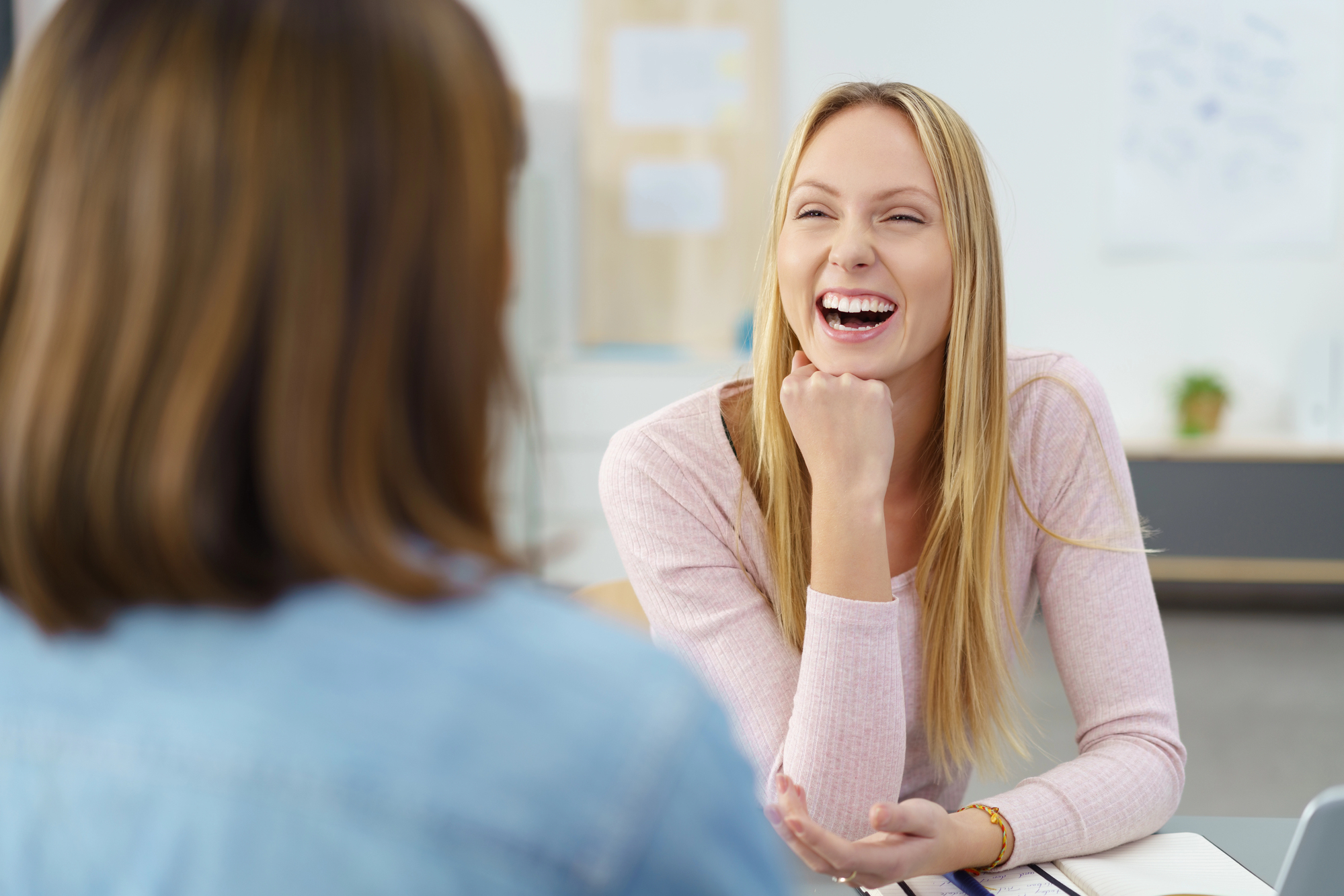 A woman with long blonde hair, wearing a light pink sweater, sits with her chin resting on her hand, smiling and laughing while engaged with another person who is out of focus in the foreground. The background is a bright, casual setting.