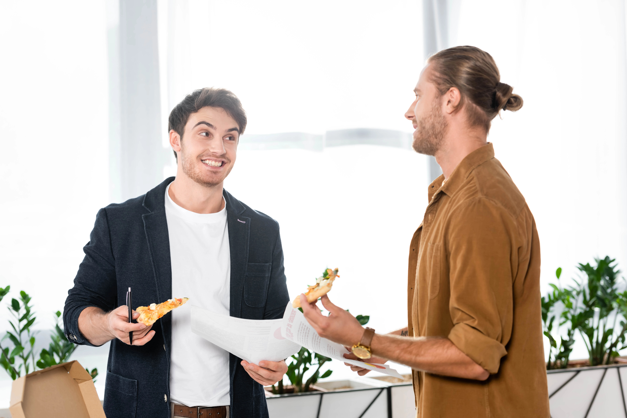 Two men are standing in an office, smiling at each other while holding slices of pizza. One is holding a document. They are near a window with potted plants in the background.