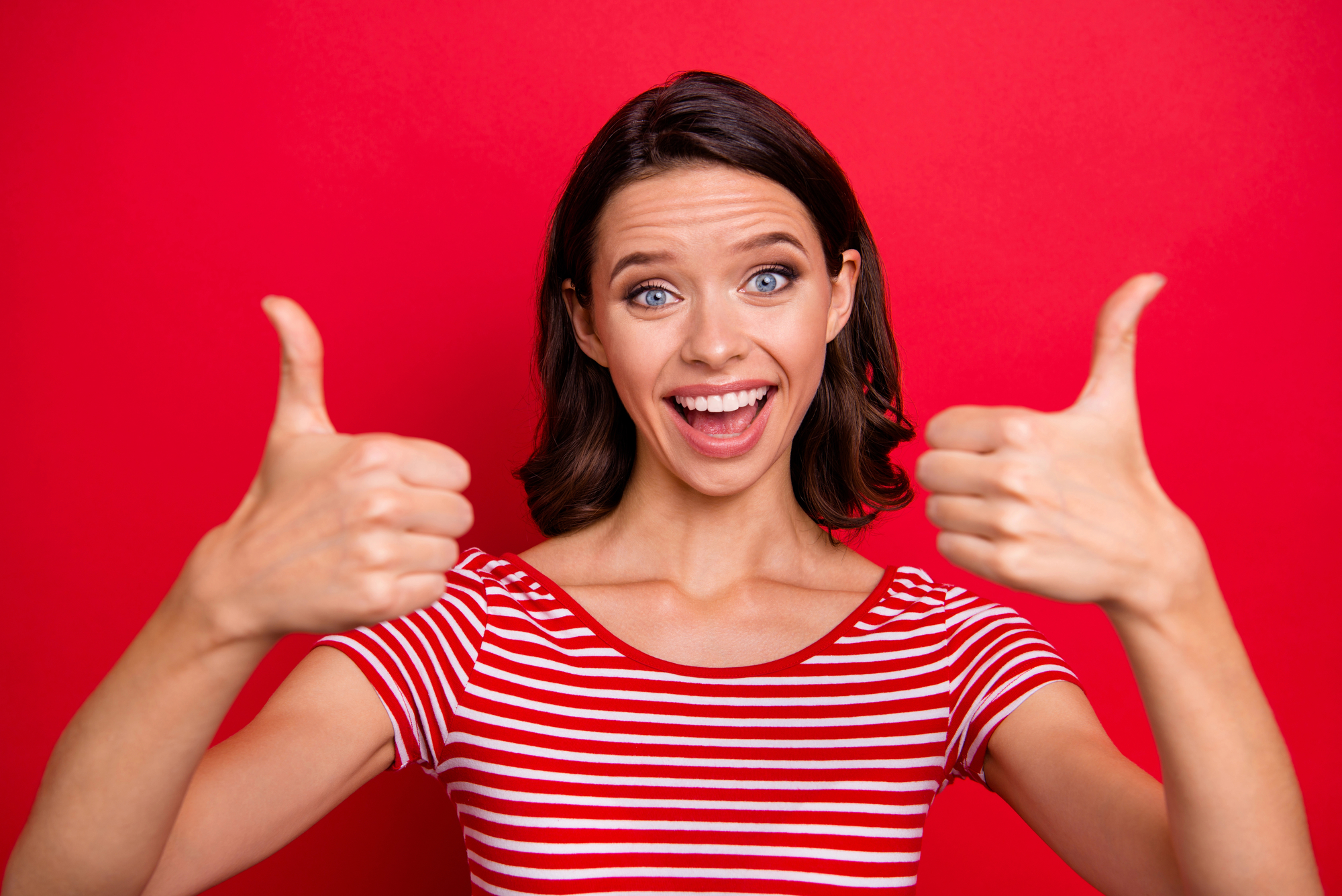 A woman with short brown hair wearing a red and white striped shirt smiles widely and gives two thumbs up in front of a bright red background.