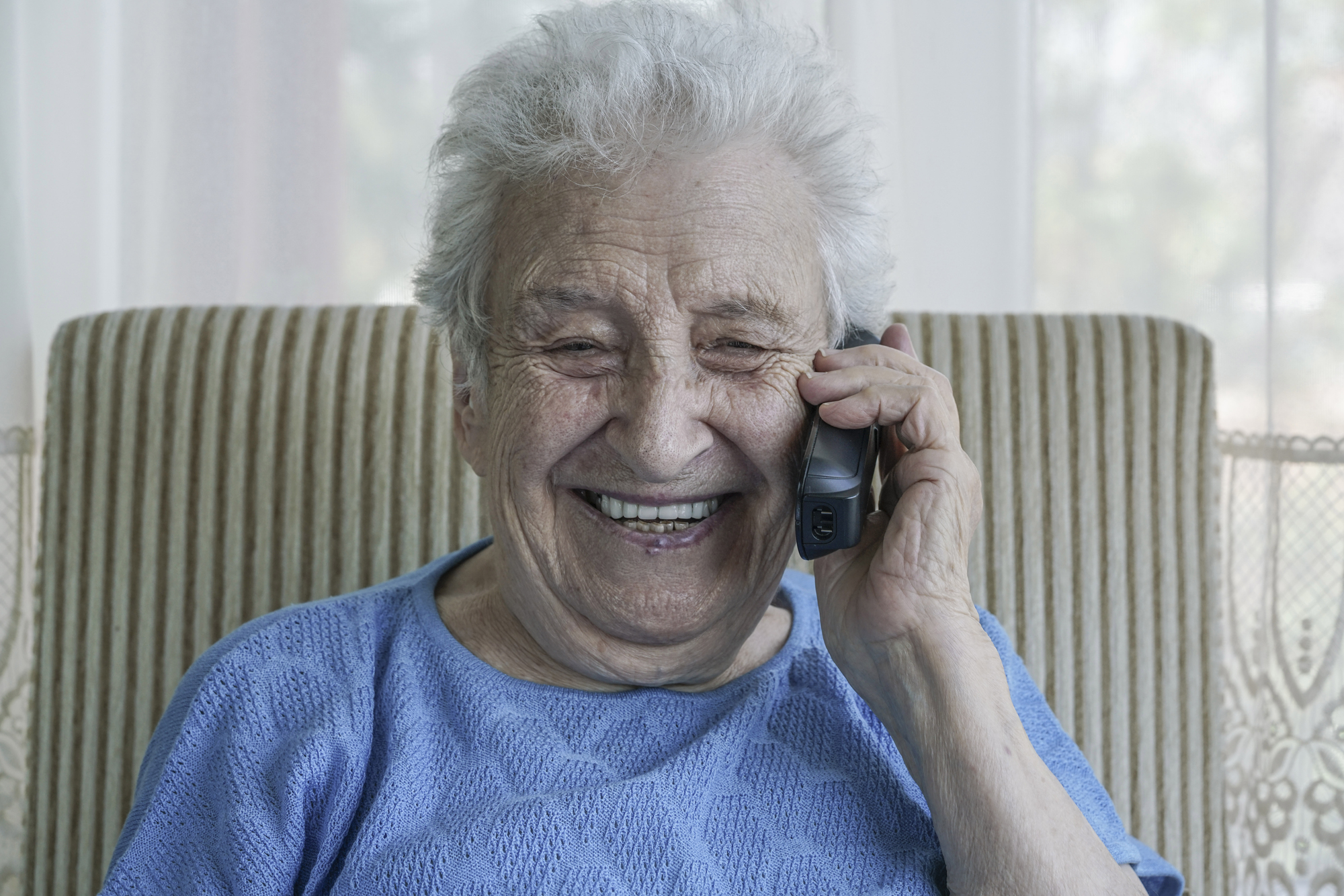 Elderly person smiling while talking on a cordless phone, sitting on a striped armchair. Wearing a light blue top, with a bright and cheerful expression in a well-lit room.