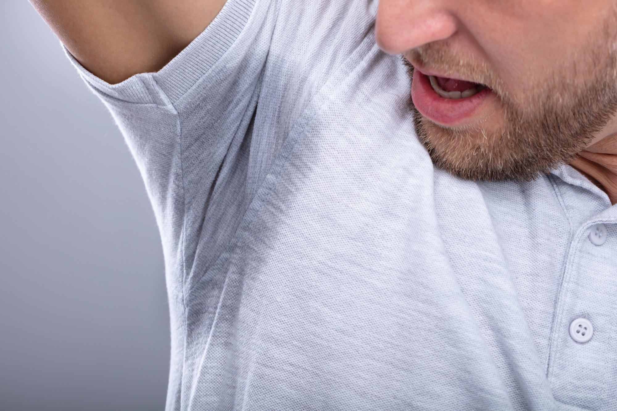 A man wearing a light gray shirt lifts his arm, revealing a large sweat stain in the armpit area. His facial expression suggests discomfort or surprise. The background is a plain gray.