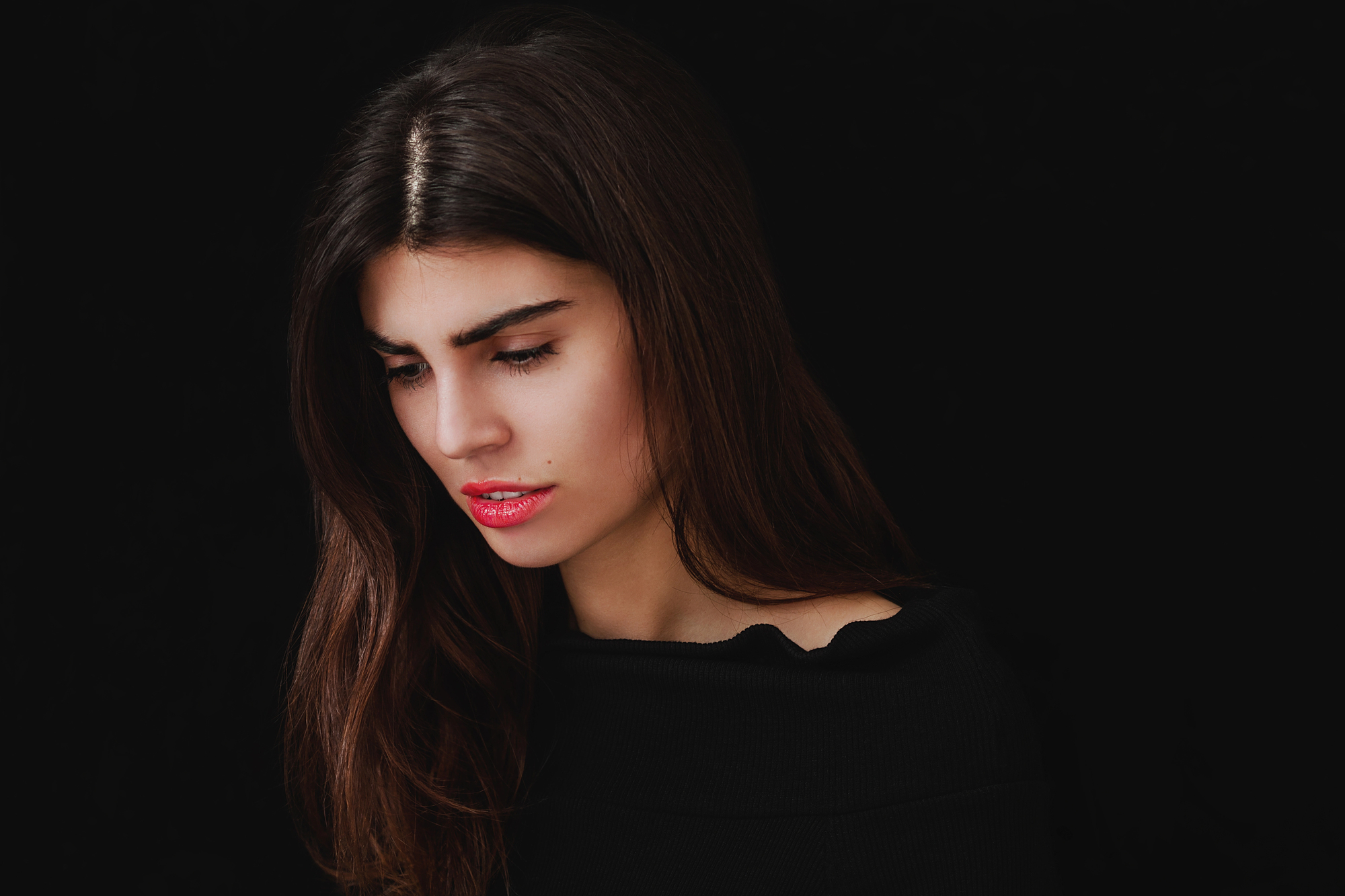 A woman with long brown hair and red lipstick looks down against a dark background. She is wearing a black top.