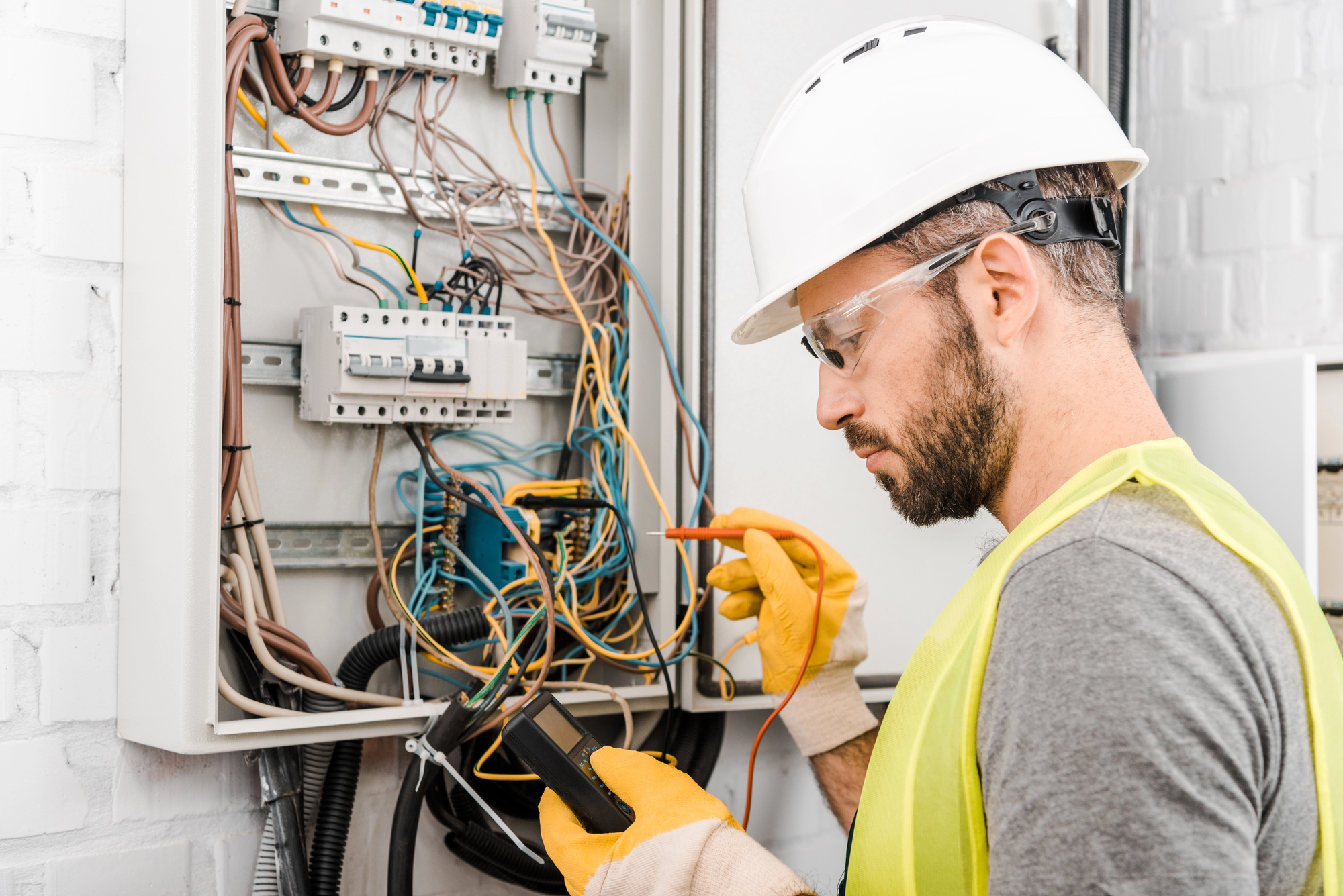 An electrician in a hard hat and safety vest uses a multimeter to inspect a circuit breaker panel. The panel is open, showing various wires and components. The electrician is wearing protective gloves and safety glasses.