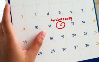 A hand holds a calendar page with the number 12 circled in red. The word "Anniversary" is written above the circled date. The calendar shows days of the month arranged in a grid.