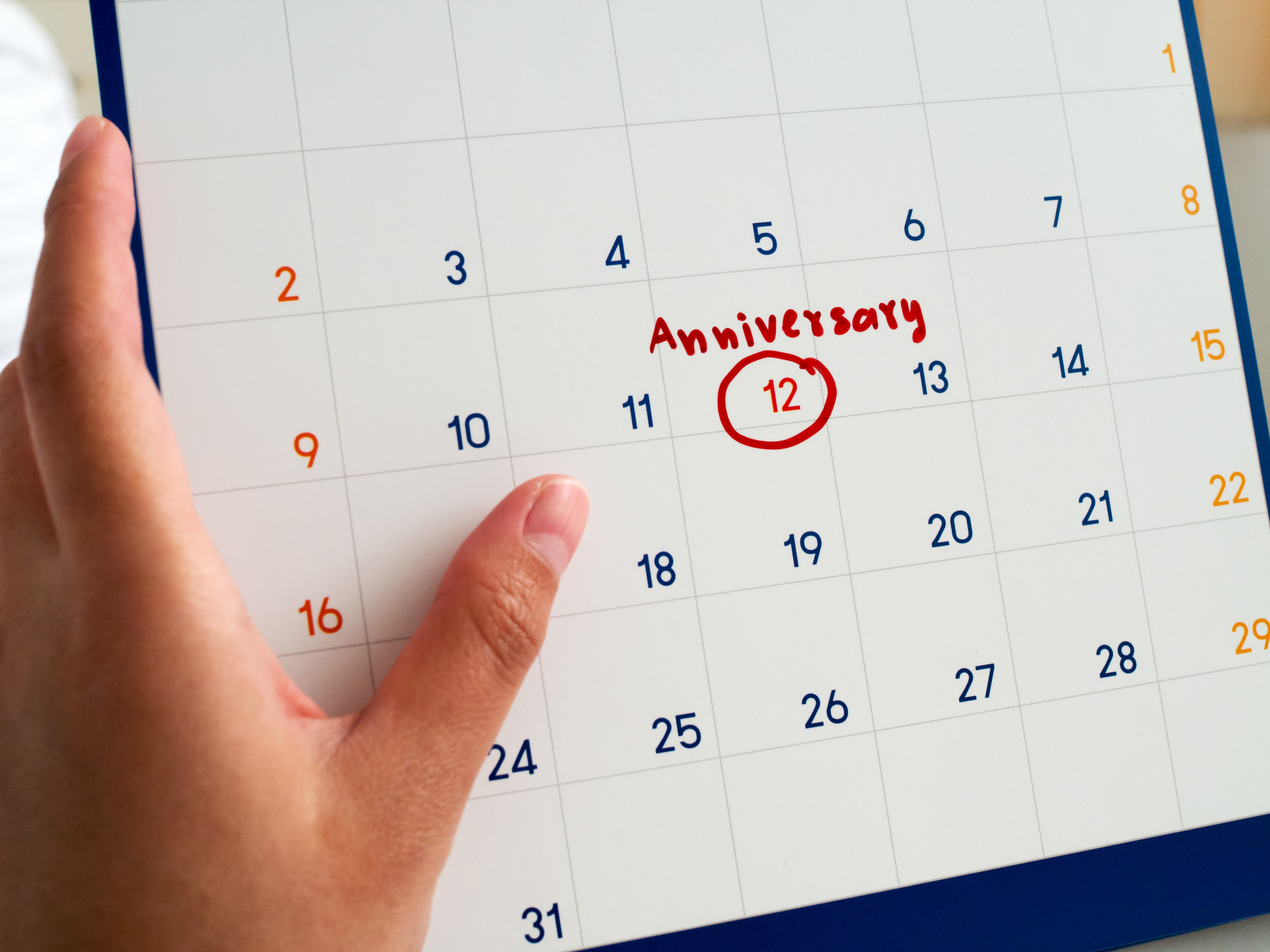 A hand holds a calendar page with the number 12 circled in red. The word "Anniversary" is written above the circled date. The calendar shows days of the month arranged in a grid.