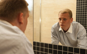 A man in a light blue shirt is intently looking at himself in a mirror in a bathroom. The wall features a mix of beige and black tiles, and the lighting is soft.