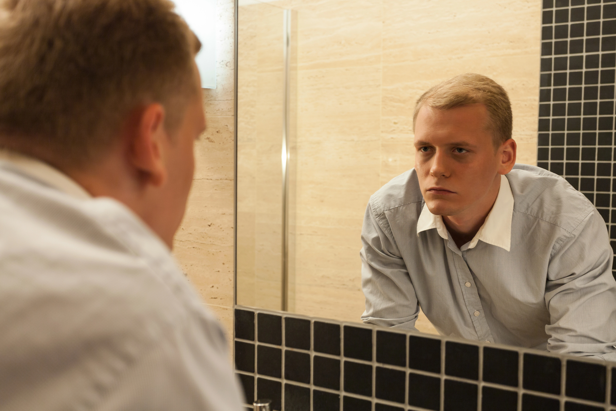 A man in a light blue shirt is intently looking at himself in a mirror in a bathroom. The wall features a mix of beige and black tiles, and the lighting is soft.