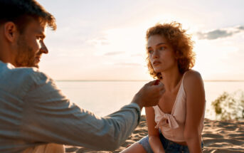 A couple enjoys a serene moment on a sunlit beach. The man offers something small to the woman, as she gazes thoughtfully towards the horizon. The background features a calm sea and a soft, glowing sky at sunset.