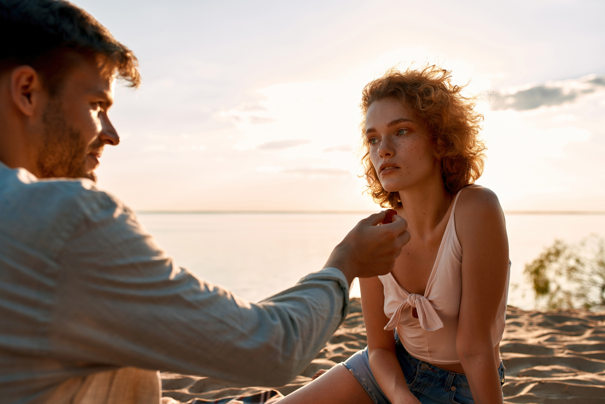 A couple enjoys a serene moment on a sunlit beach. The man offers something small to the woman, as she gazes thoughtfully towards the horizon. The background features a calm sea and a soft, glowing sky at sunset.