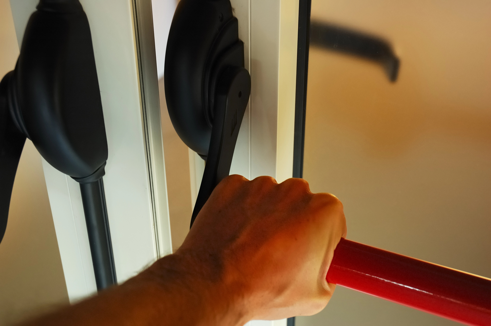 A person grips a red metal bar, opening a black lever handle on a door. The scene is indoors, highlighting the hand's action on the door mechanism, suggesting the door is being pushed or secured.