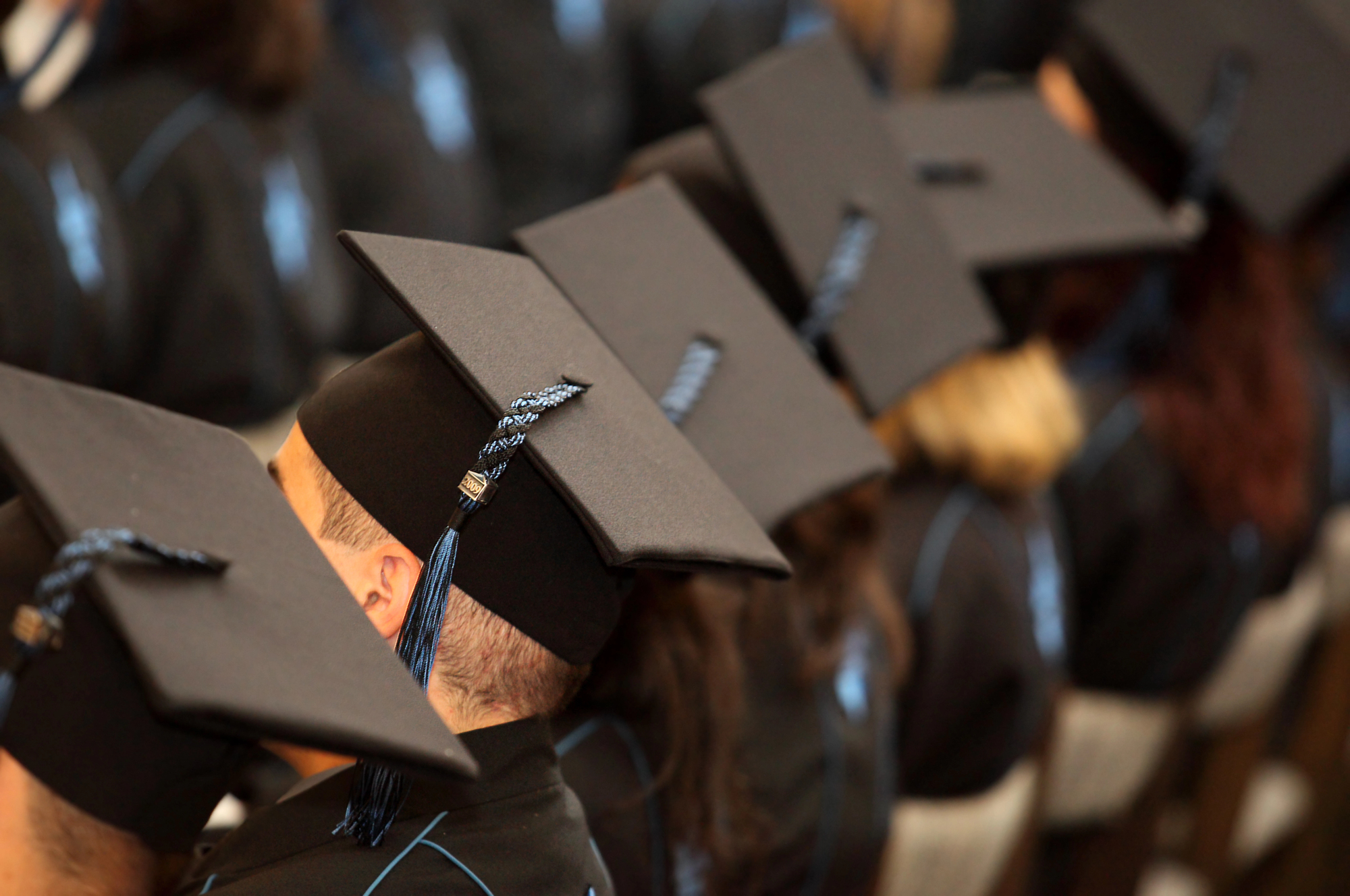 Graduates in black caps and gowns sit in a row, focusing forward. Their tassels hang on the right side, suggesting a ceremony in progress. The image captures a moment of formal academic celebration.