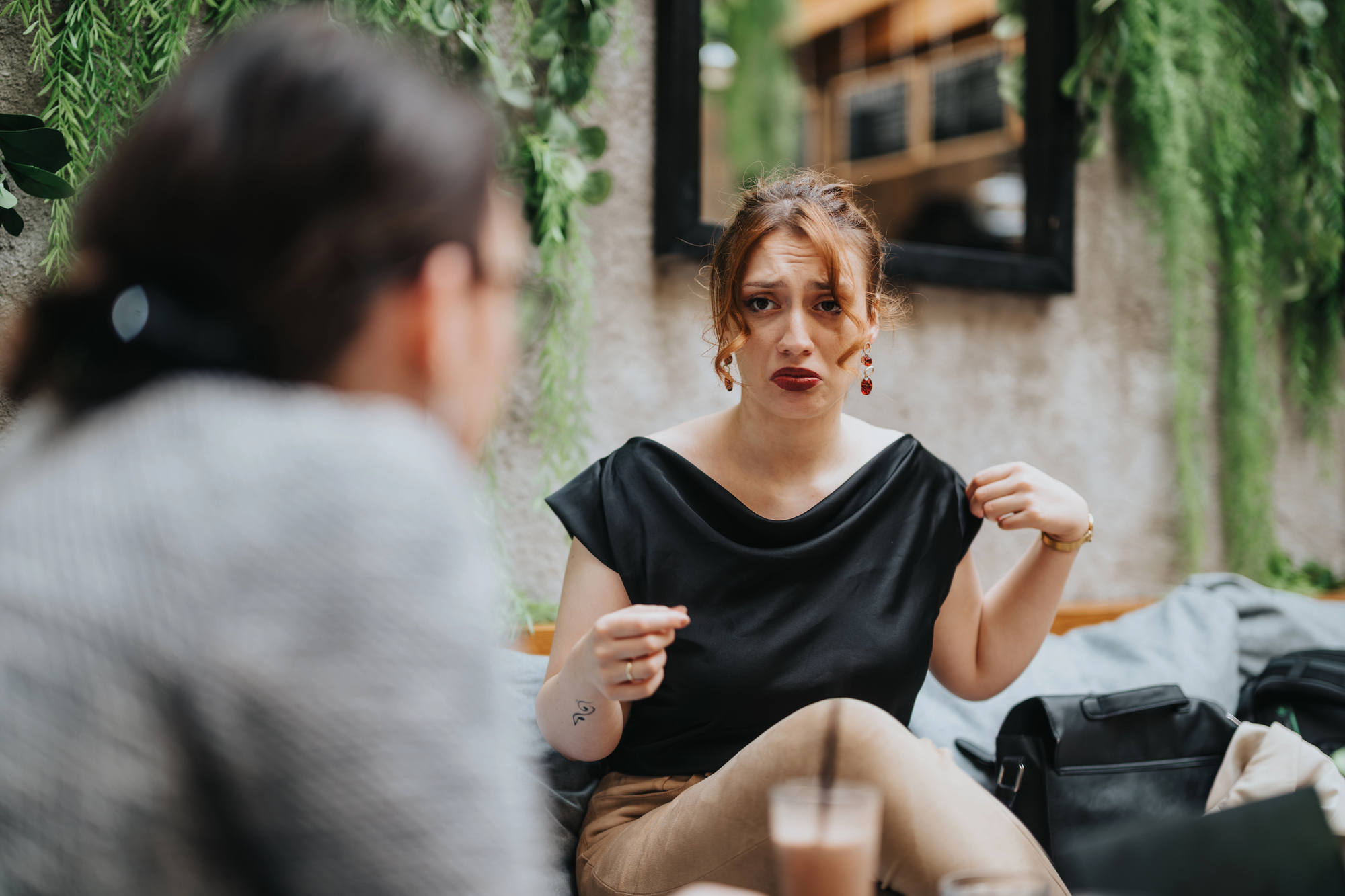 A woman in a black top sits at a table in a cafe, looking distressed, while talking to someone. She gestures with her hands, surrounded by a green, leafy decor. A drink is on the table.