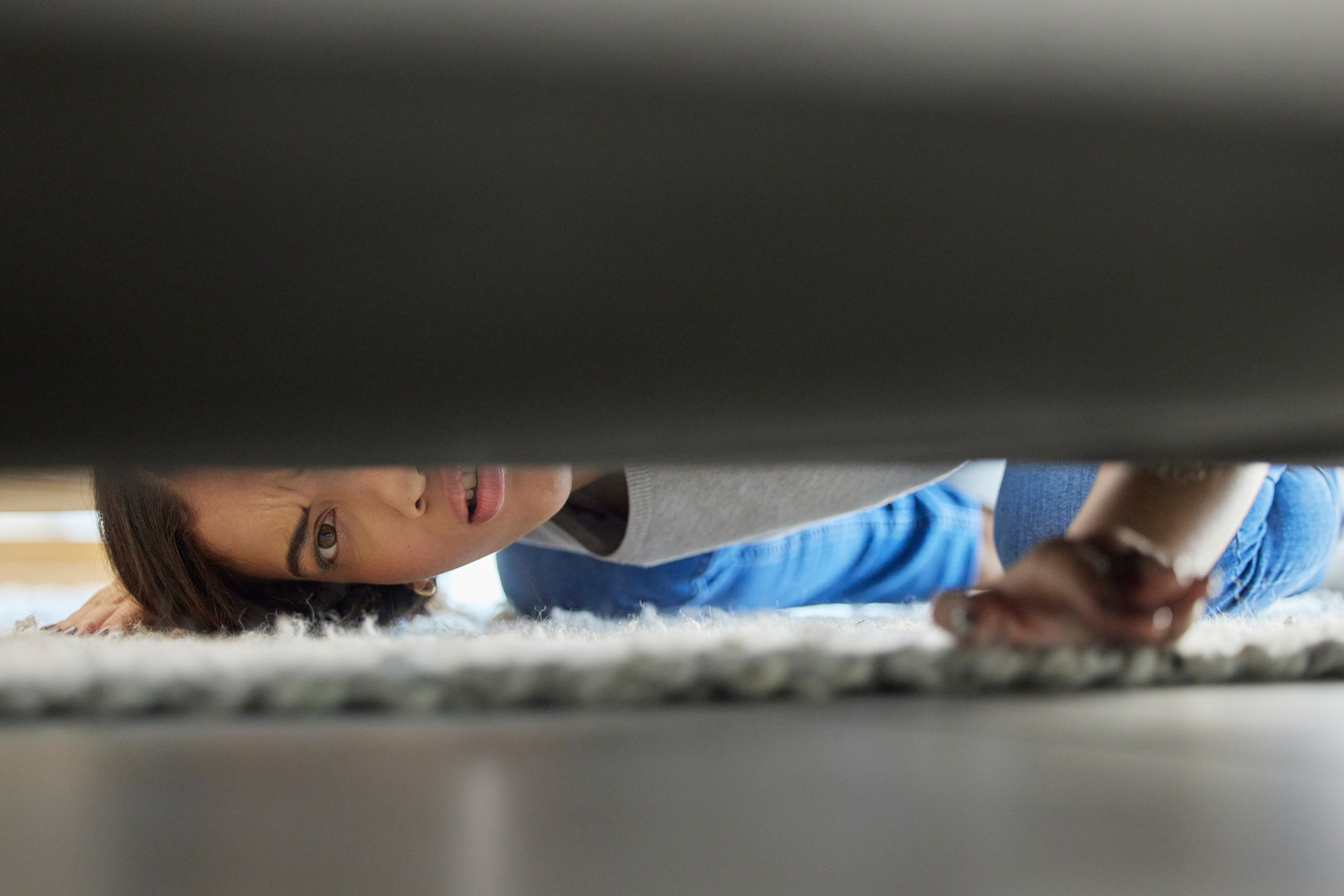 A woman lying on the floor peeks under a sofa, looking concerned or searching for something. She is wearing a casual outfit and is partially visible behind the sofa, with her hand reaching underneath.