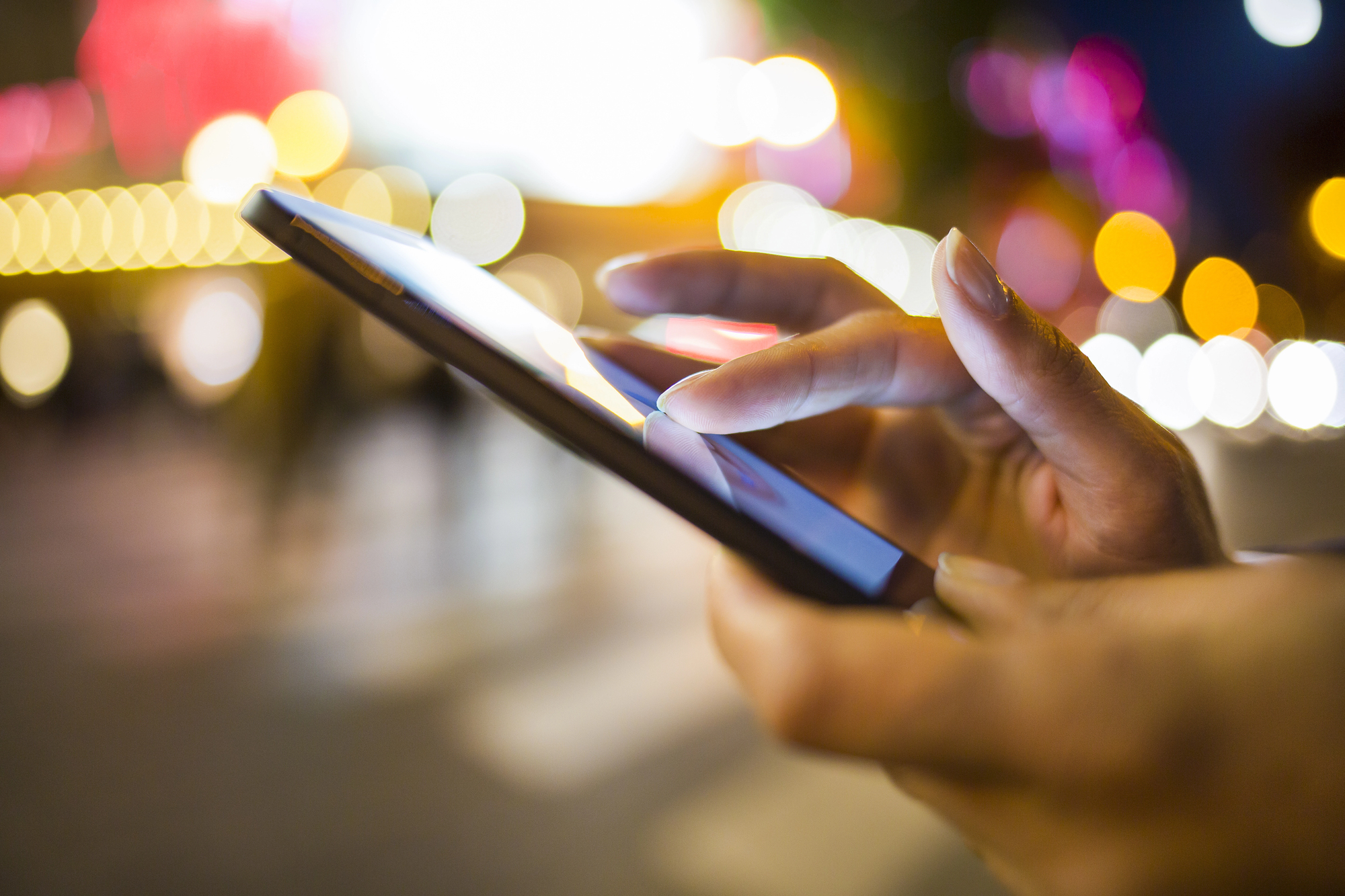 Close-up of a person's hands using a smartphone at night, with blurred city lights in the background. The bright bokeh effect creates a vibrant atmosphere.