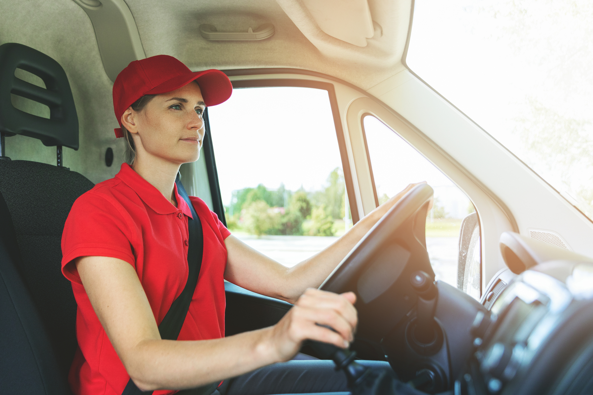 A person wearing a red cap and shirt is driving a vehicle, focused on the road. Sunlight streams through the window, illuminating the interior. The driver is seated in a modern van, holding the steering wheel with both hands.