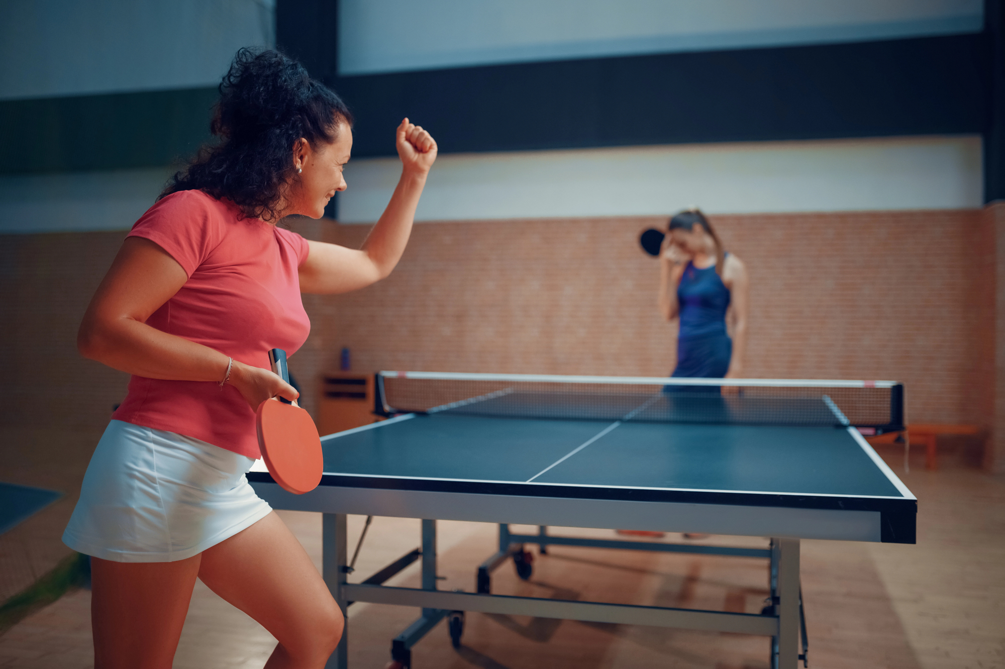 A woman in a pink shirt and white shorts celebrates with her fist raised, standing next to a ping pong table. Another woman in a blue dress holds her head in disappointment on the opposite side of the table.