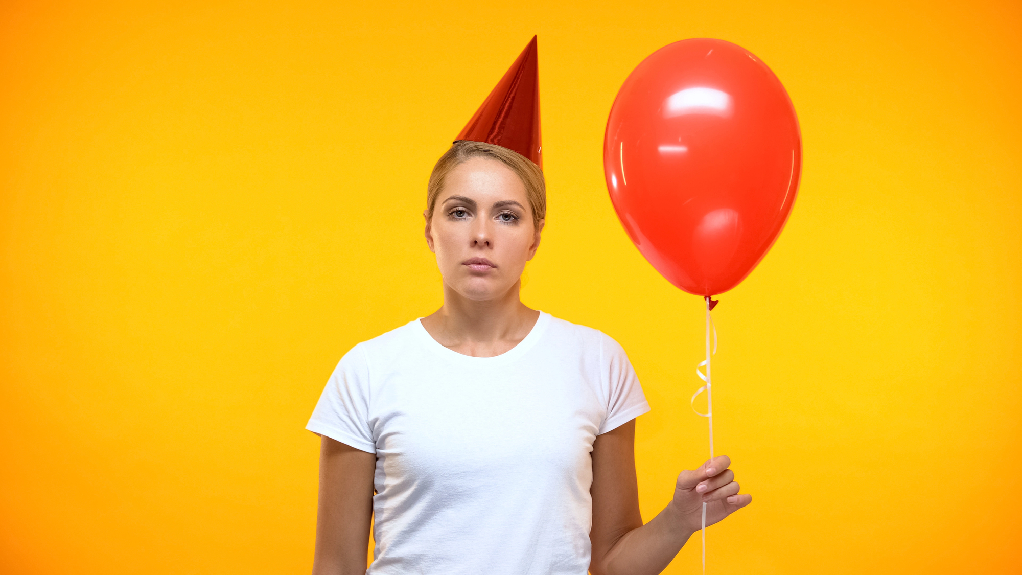 A woman in a white t-shirt stands against a bright yellow background. She wears a red party hat and holds a red balloon, looking serious.