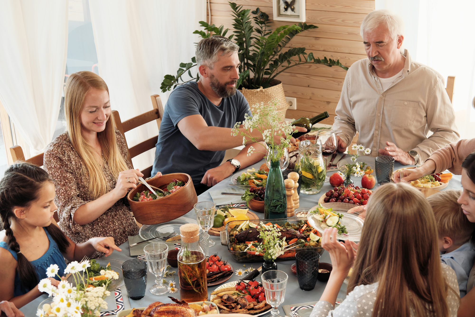 A family gathered around a dining table enjoying a meal. The table is filled with various dishes, including fruits, salads, and drinks. The room is decorated with plants and natural light streams through a window. People are engaged in conversation.