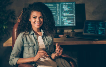 A person with curly hair sits in front of a desk with two monitors displaying code. They are holding glasses and smiling, wearing a denim jacket over a white shirt. The room has a relaxed, dimly lit ambiance.