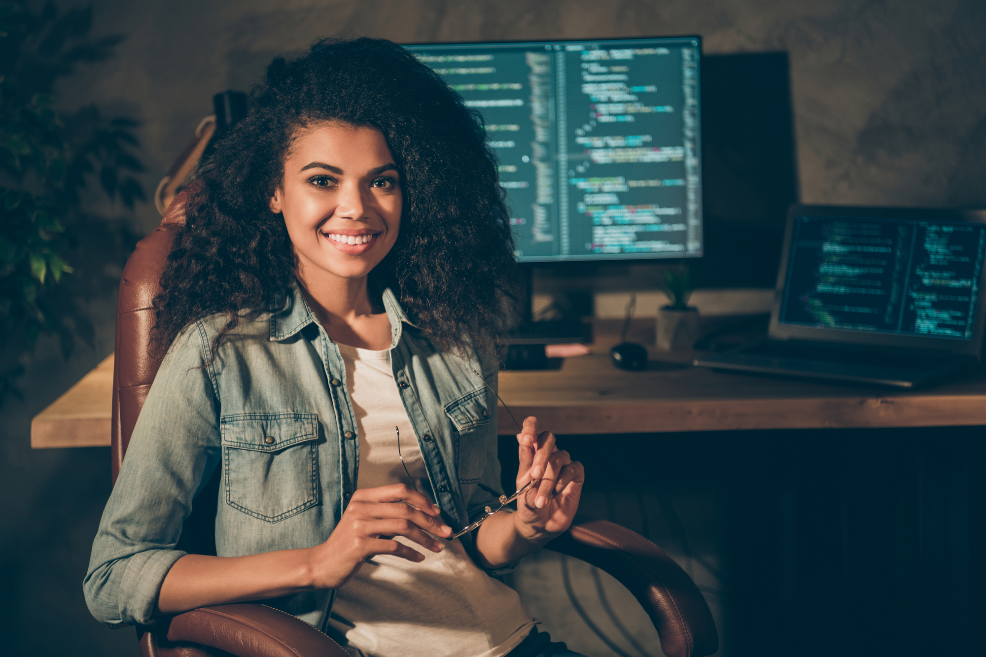 A person with curly hair sits in front of a desk with two monitors displaying code. They are holding glasses and smiling, wearing a denim jacket over a white shirt. The room has a relaxed, dimly lit ambiance.