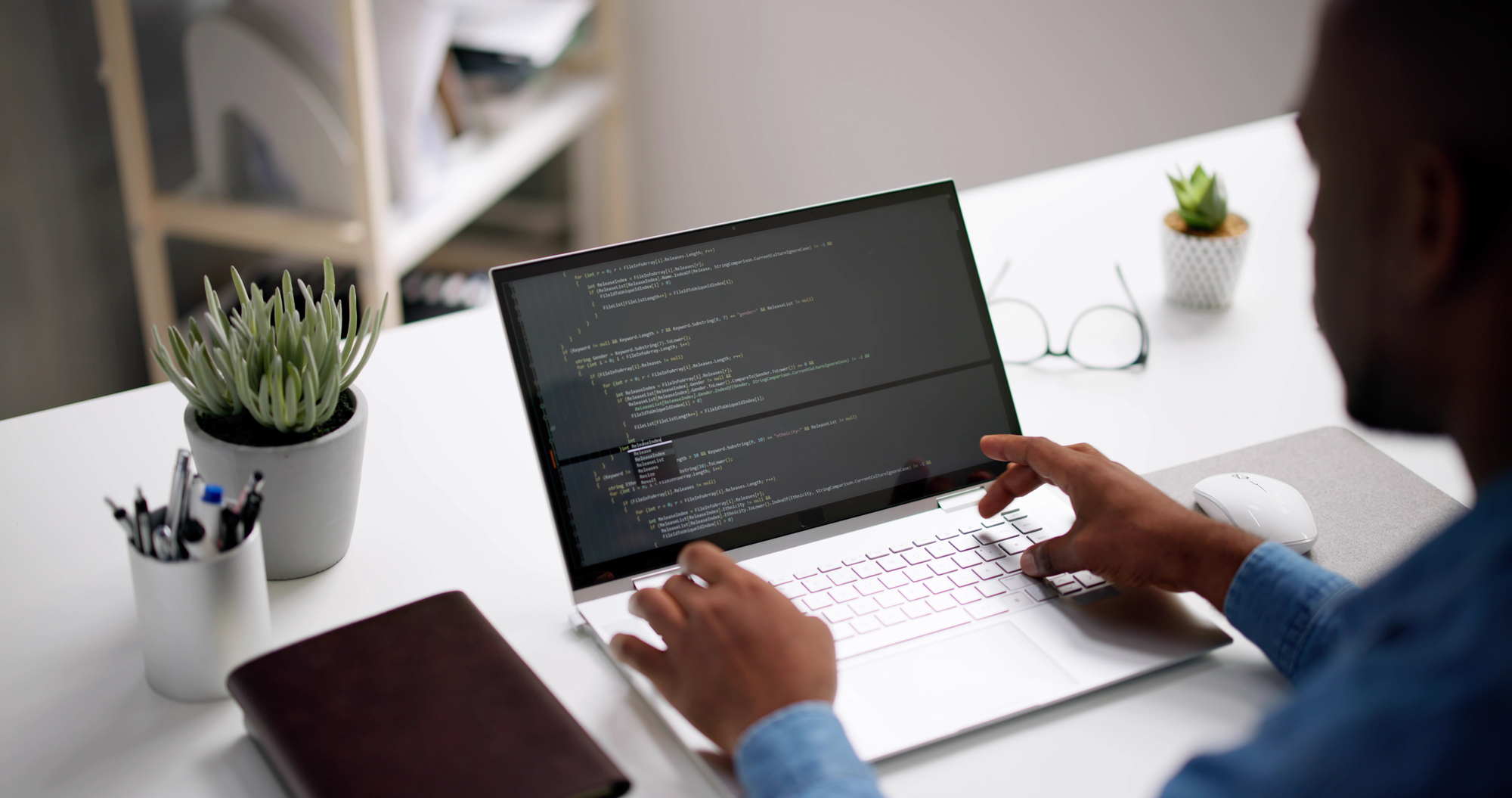 A person is typing code on a laptop at a desk. The screen displays lines of programming code. Nearby are a potted plant, a closed notebook, and a pair of glasses. The workspace has a clean, organized look.