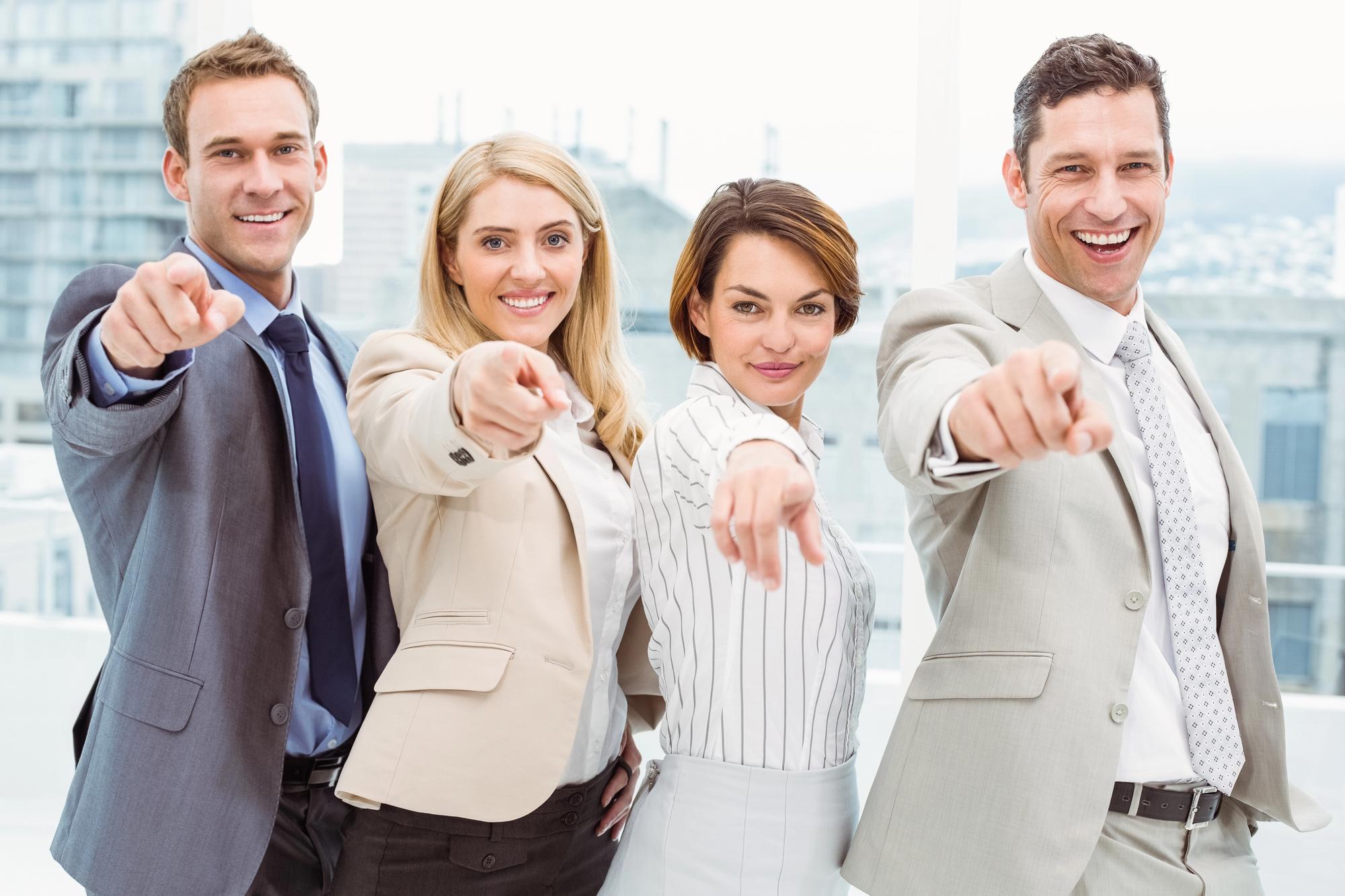 Four people in business attire stand indoors, smiling and pointing forward. The group includes two men and two women, all looking confident and positive, with a bright office background.