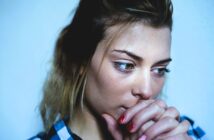 A woman with light brown hair gazes downward thoughtfully, resting her chin on her clasped hands. She wears a checkered shirt and has red nails, set against a plain blue background.