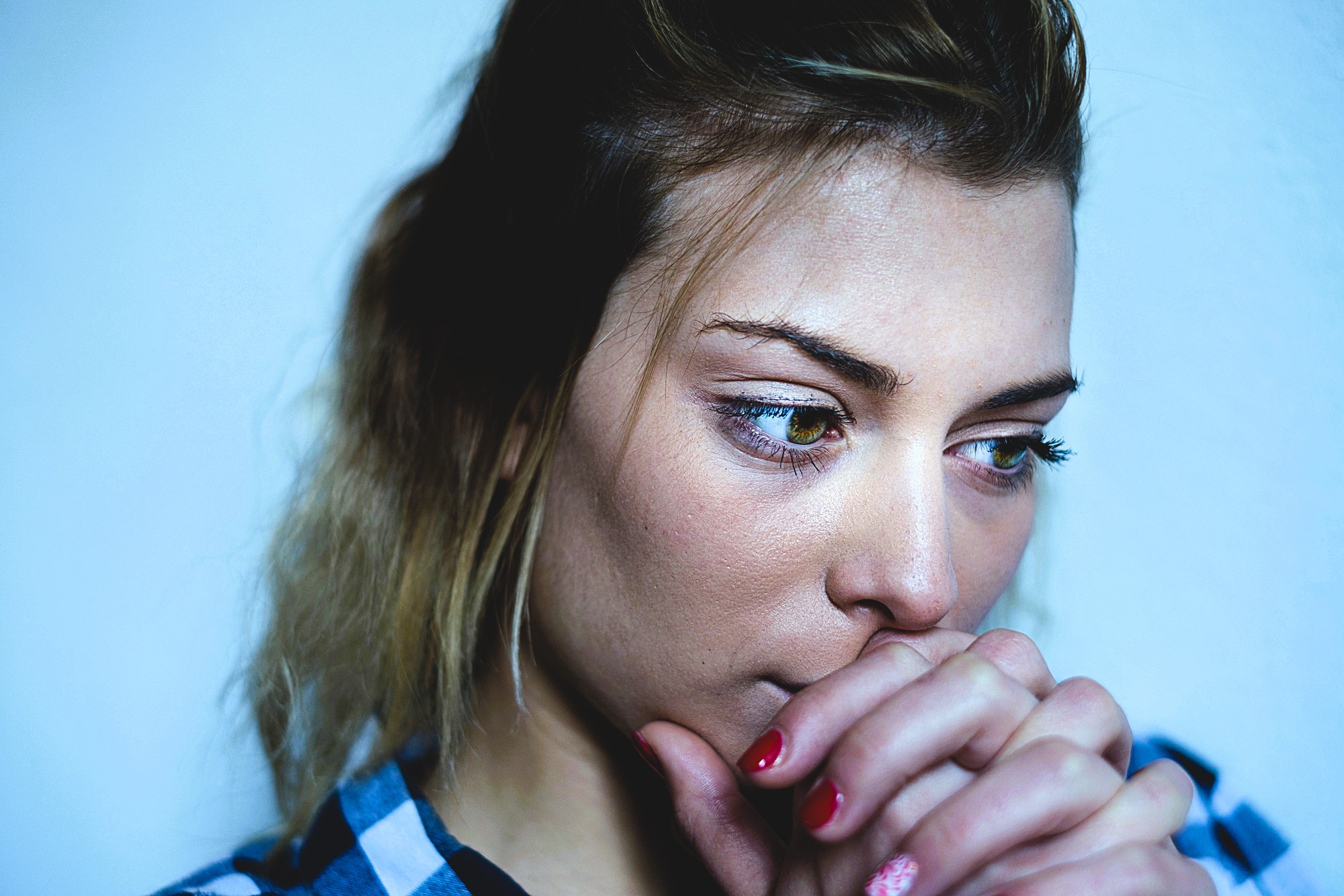 A woman with light brown hair gazes downward thoughtfully, resting her chin on her clasped hands. She wears a checkered shirt and has red nails, set against a plain blue background.