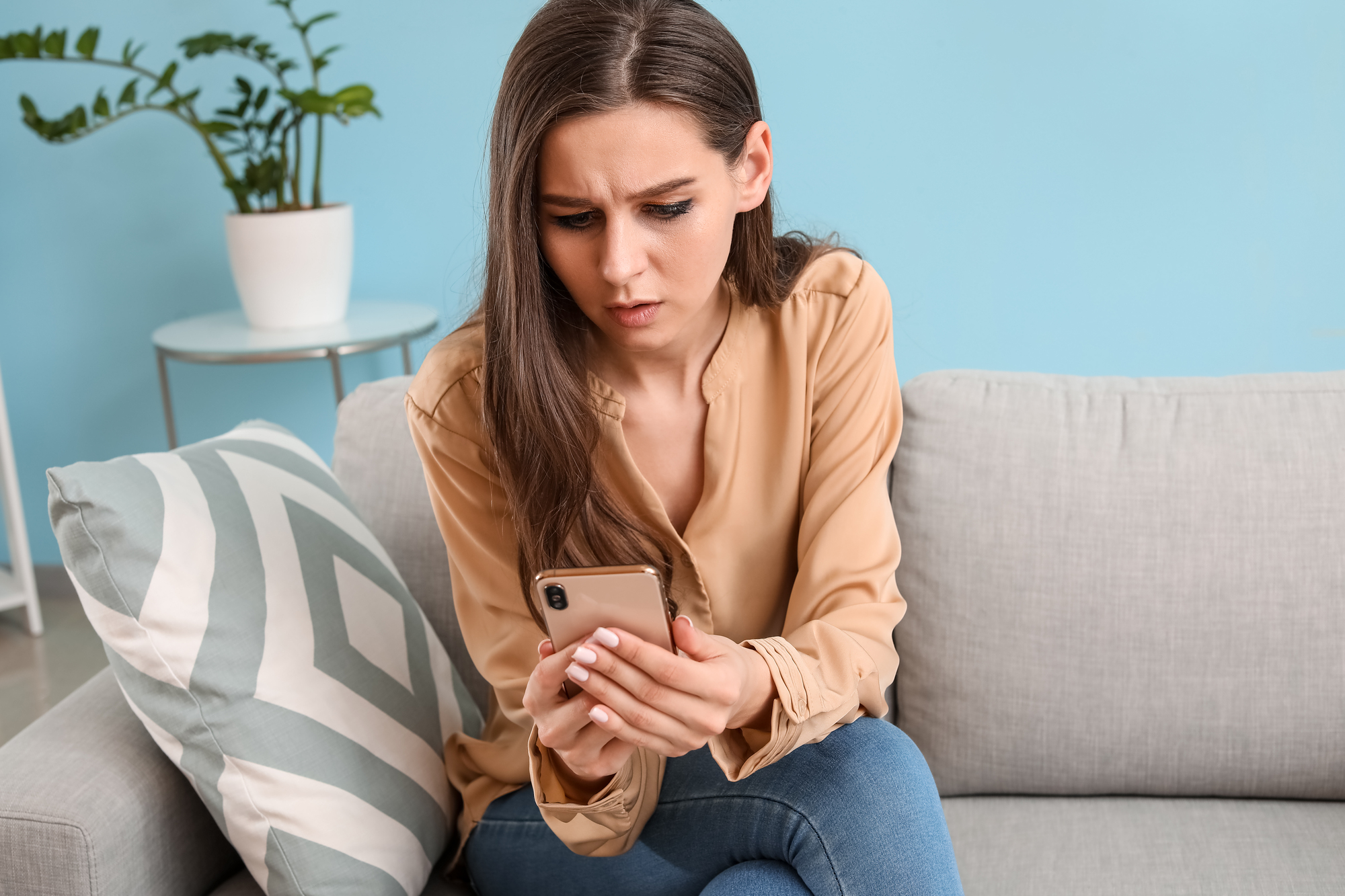 A woman with long brown hair, wearing a beige blouse and jeans, looks at her phone with a concerned expression. She sits on a light gray sofa with a patterned pillow and a potted plant in the background.