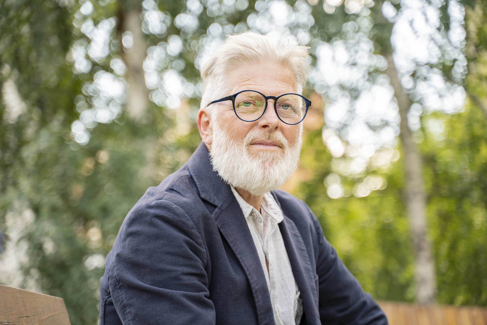 An older man with white hair and a beard, wearing glasses, a navy blazer, and a light shirt, sits on a bench outdoors with trees in the background.