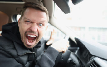 A man with a beard sits in a car, gripping the steering wheel, and looking at the camera with an open mouth and wide eyes, appearing agitated or frustrated. He wears a black jacket and has a short haircut.