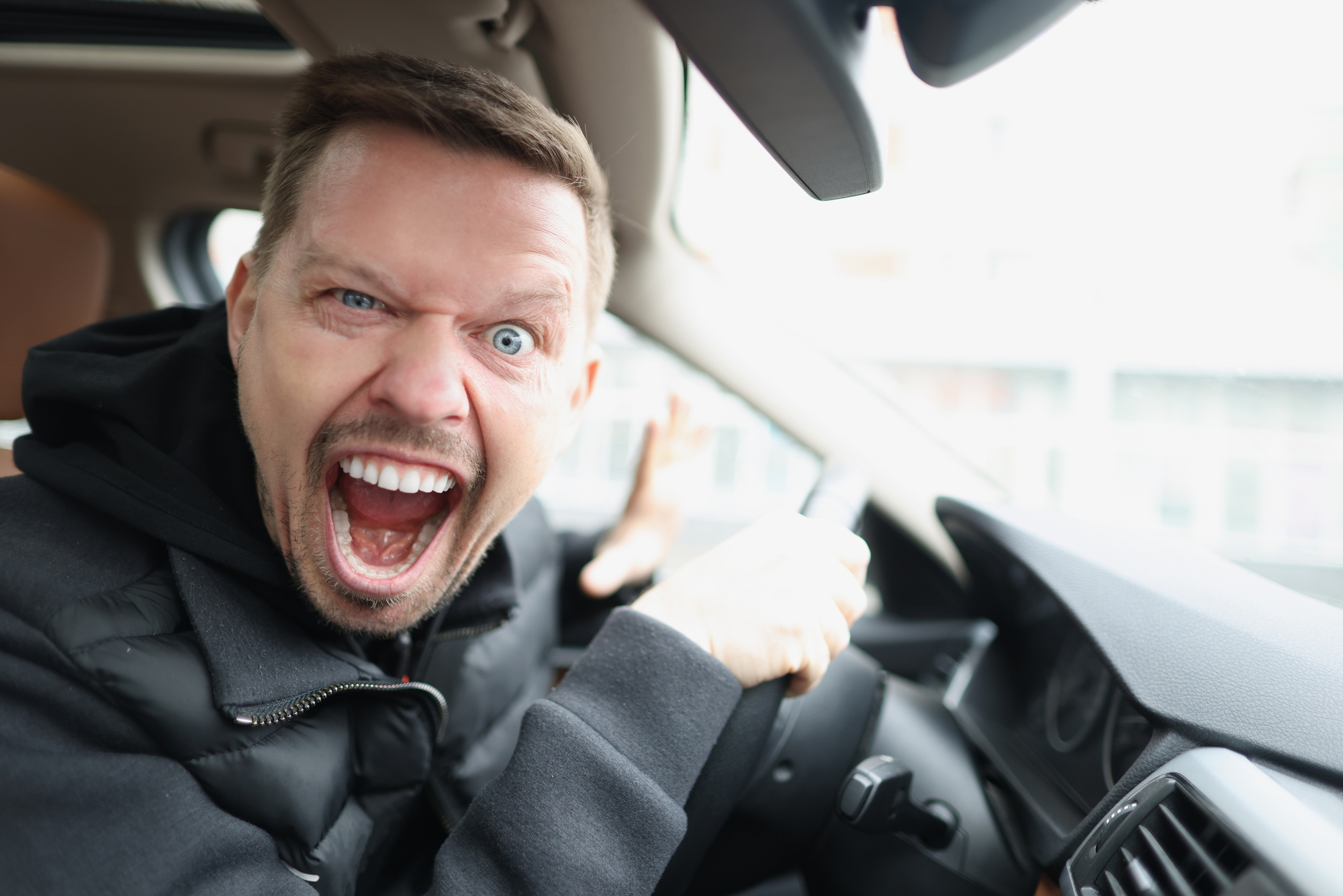 A man with a beard sits in a car, gripping the steering wheel, and looking at the camera with an open mouth and wide eyes, appearing agitated or frustrated. He wears a black jacket and has a short haircut.