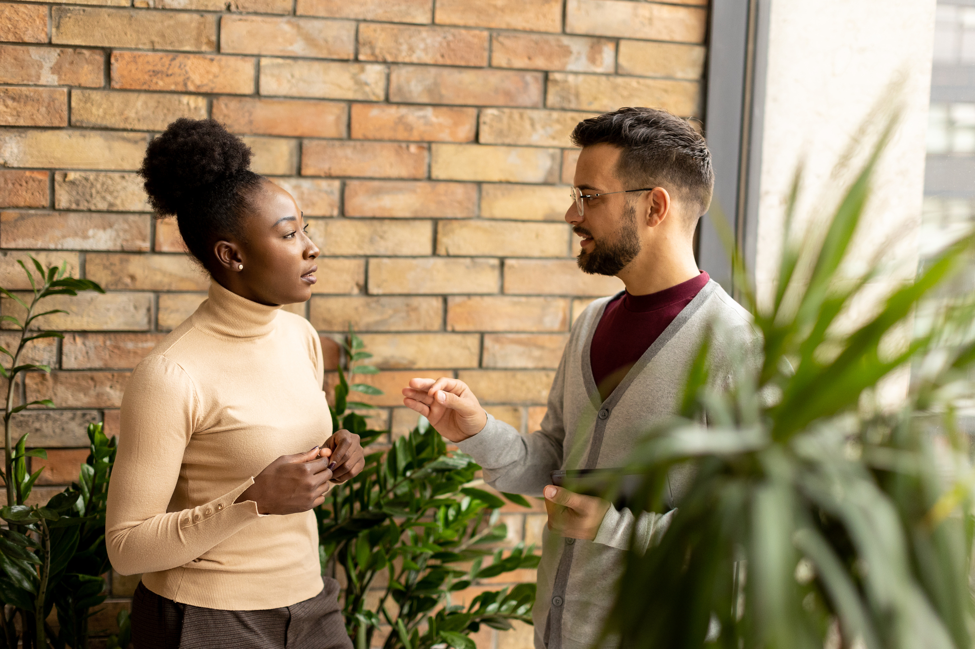 A man and woman are engaged in conversation indoors. The woman, wearing a beige turtleneck, faces the man, who wears glasses and a gray sweater. They stand by a brick wall, surrounded by potted plants. Natural light streams through a nearby window.