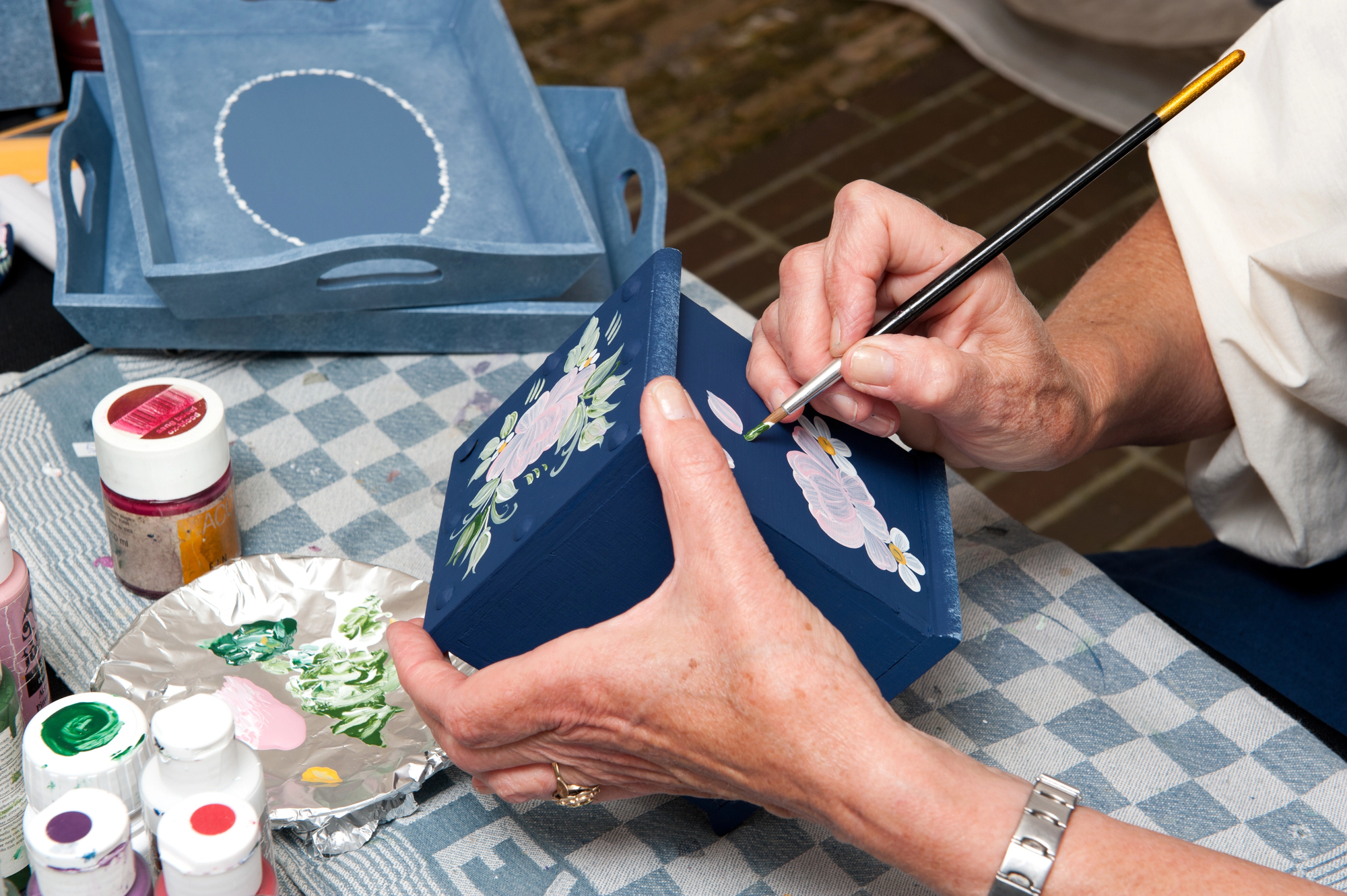 A person is painting a dark blue box with floral designs while holding a paintbrush. Nearby are bottles of paint and a paint palette with various colors. The background features a patterned tablecloth.