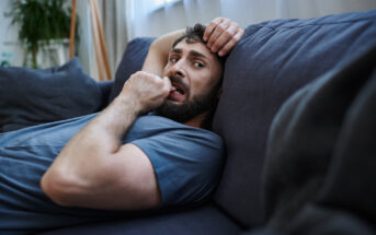 A man with a beard lounges on a dark sofa, gazing at the camera. He wears a blue T-shirt and appears to be biting his fingernail, with one hand resting on his head. A large window and a plant are visible in the background.