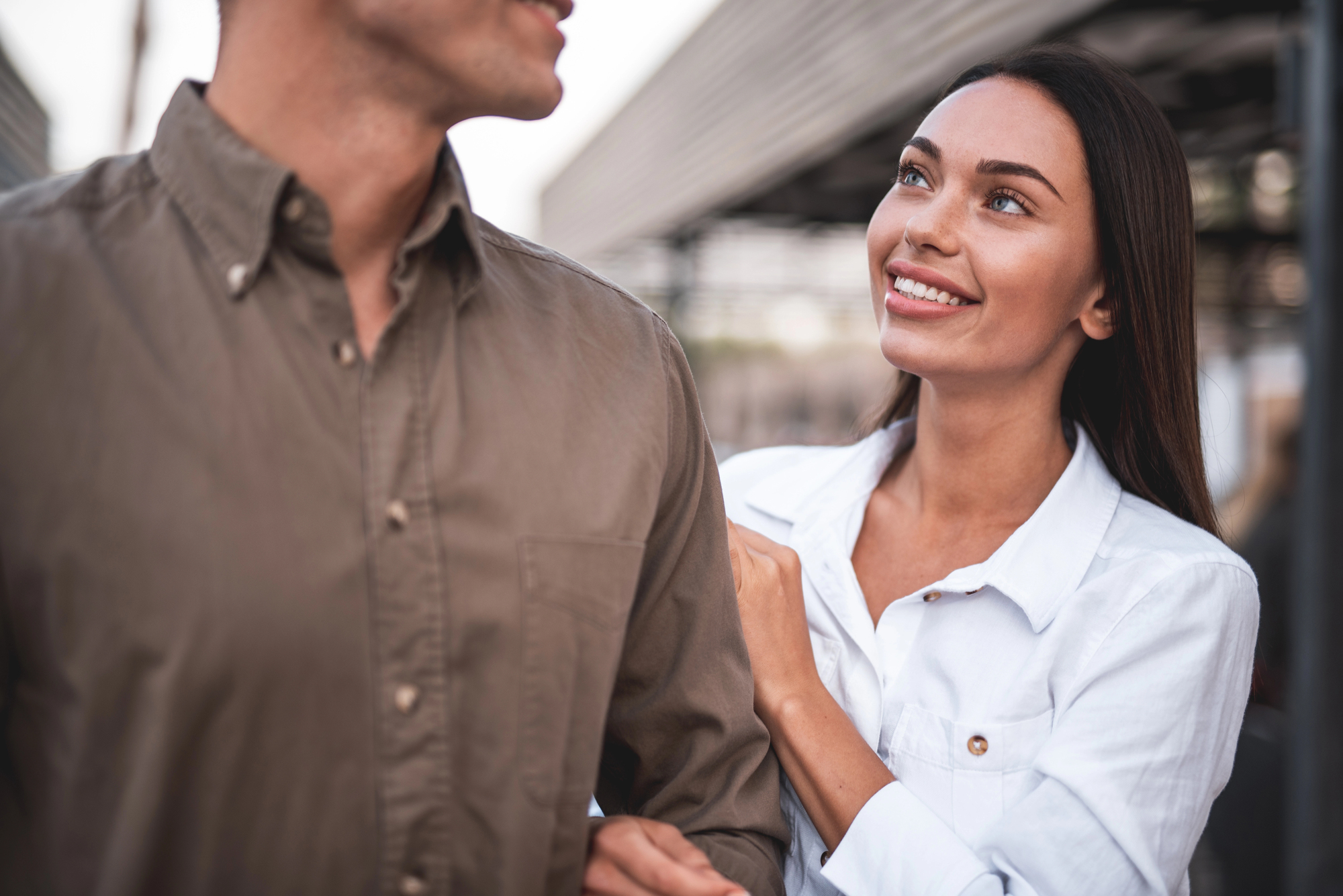 A woman in a white shirt smiles while looking at a man in a brown shirt. She gently touches his shoulder with her hand. They are outdoors, with blurred buildings in the background.