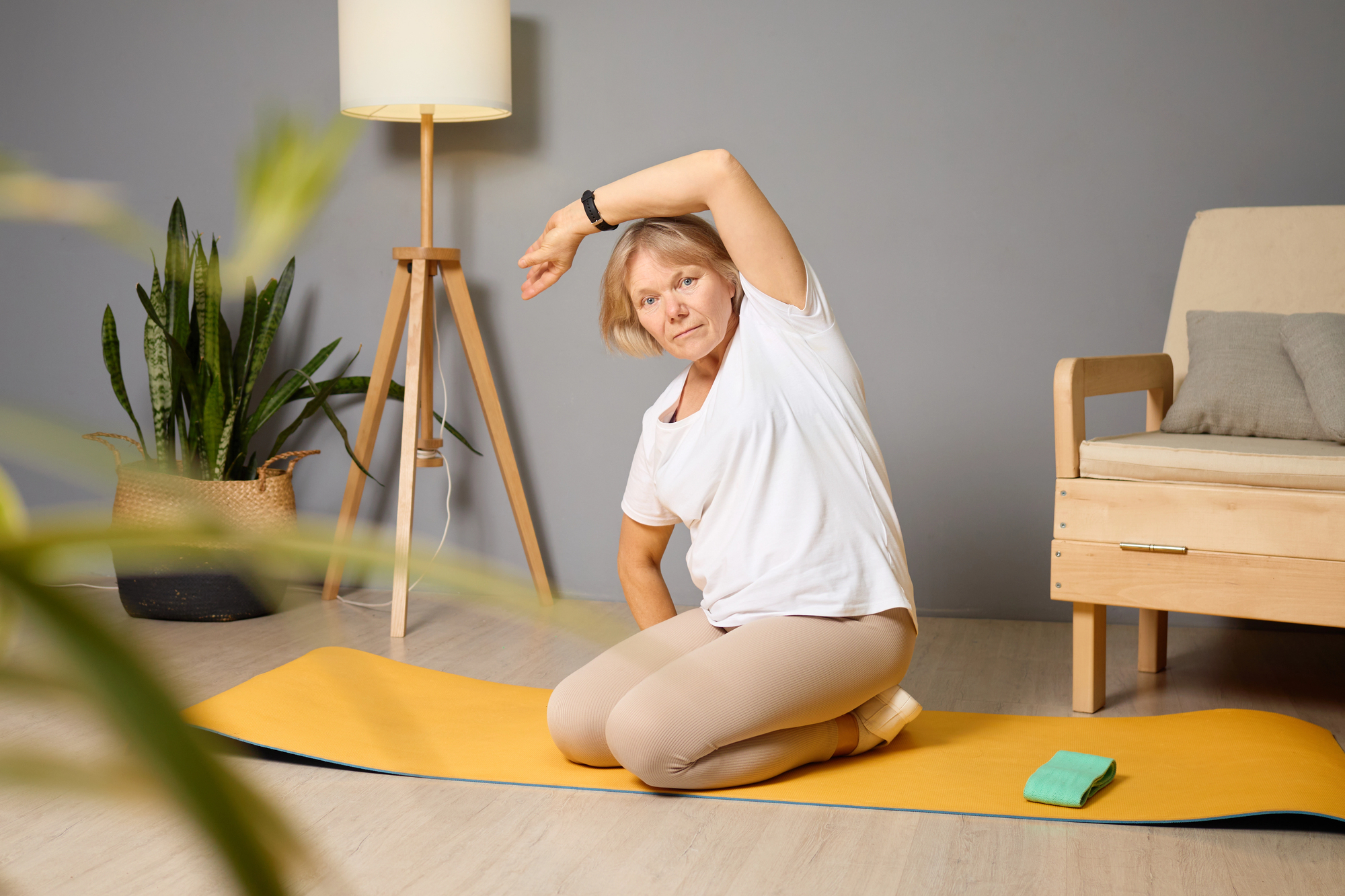 An older woman in a white shirt and beige pants sits on a yellow yoga mat, performing a side stretch with one arm over her head. She is in a cozy room with a potted plant, a standing lamp, and a wooden chair in the background.