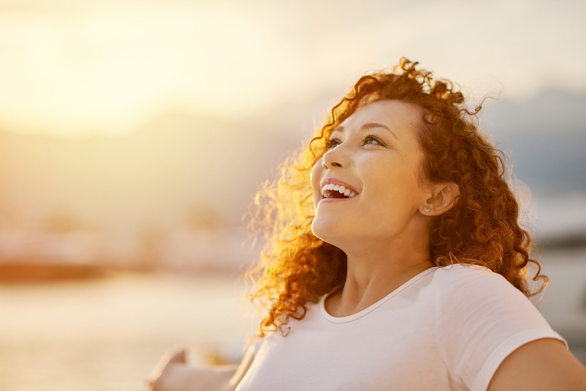 A woman with curly red hair and a white shirt is smiling while looking upwards. She is outdoors, with a blurred background of mountains and water, bathed in warm, golden sunlight.