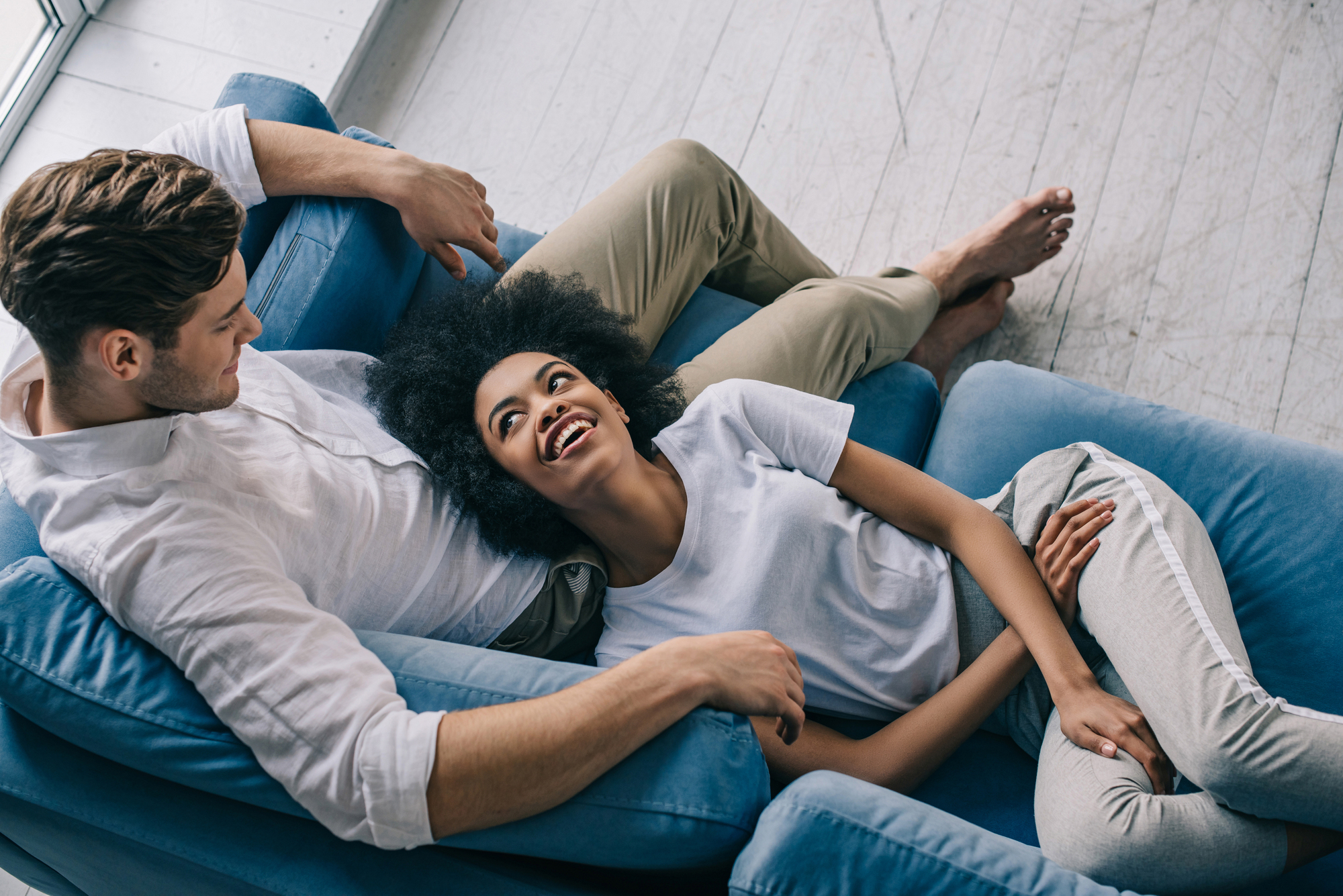 A man and woman are lounging comfortably on a blue sofa. The woman is lying across the man's lap, and they are both smiling and looking at each other. They appear relaxed in a bright room with a wooden floor.