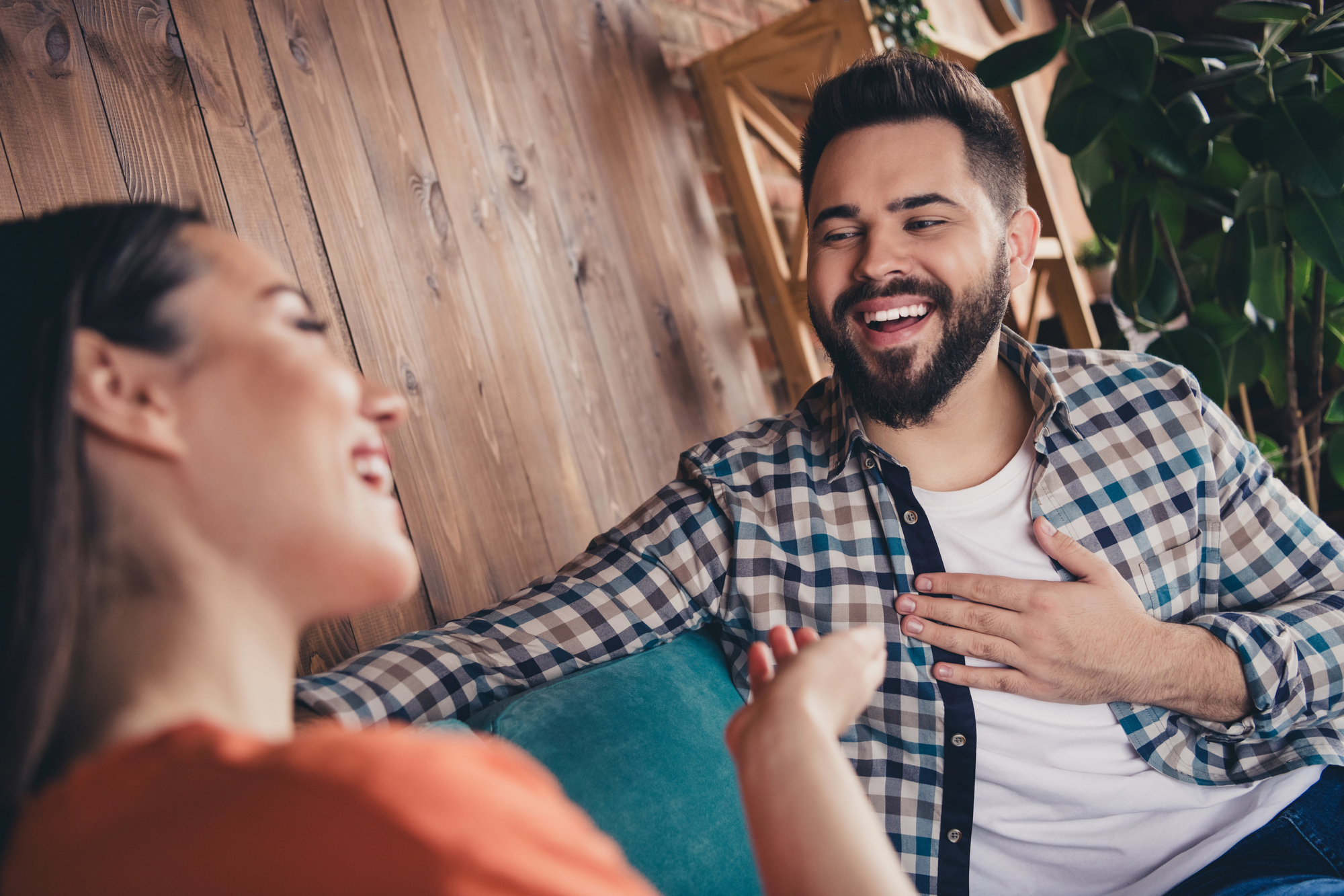 A man and woman are sitting on a couch, engaged in a lively conversation. The man, with a beard and wearing a checkered shirt, is smiling and gesturing with his hand on his chest. The woman wears an orange top and faces him, also smiling.