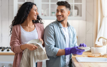 A man and woman are smiling at each other while washing dishes in a bright kitchen. The man wears purple gloves and holds a plate, while the woman holds a towel and another plate. Kitchen shelves with dishes and utensils are visible in the background.