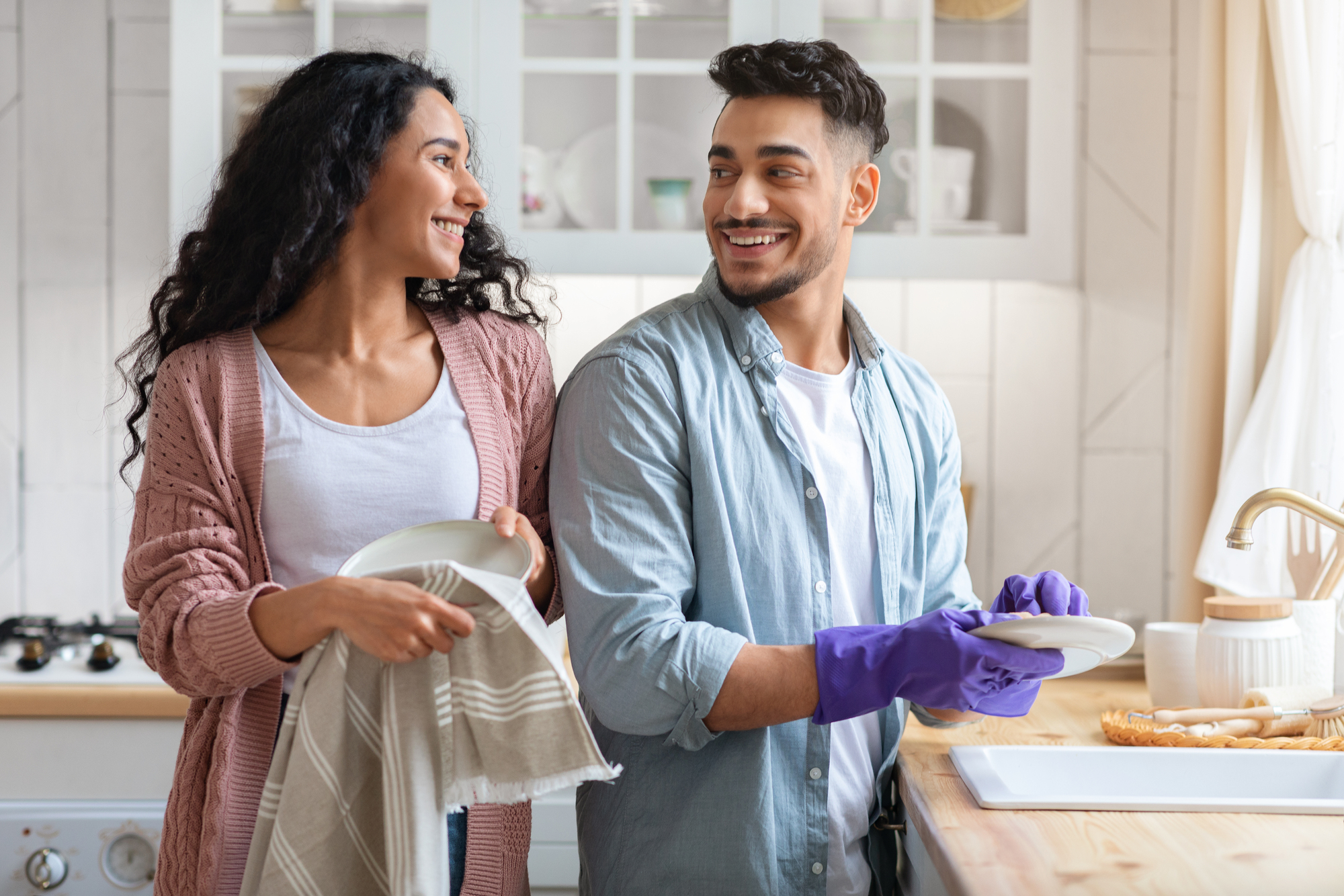 A man and woman are smiling at each other while washing dishes in a bright kitchen. The man wears purple gloves and holds a plate, while the woman holds a towel and another plate. Kitchen shelves with dishes and utensils are visible in the background.