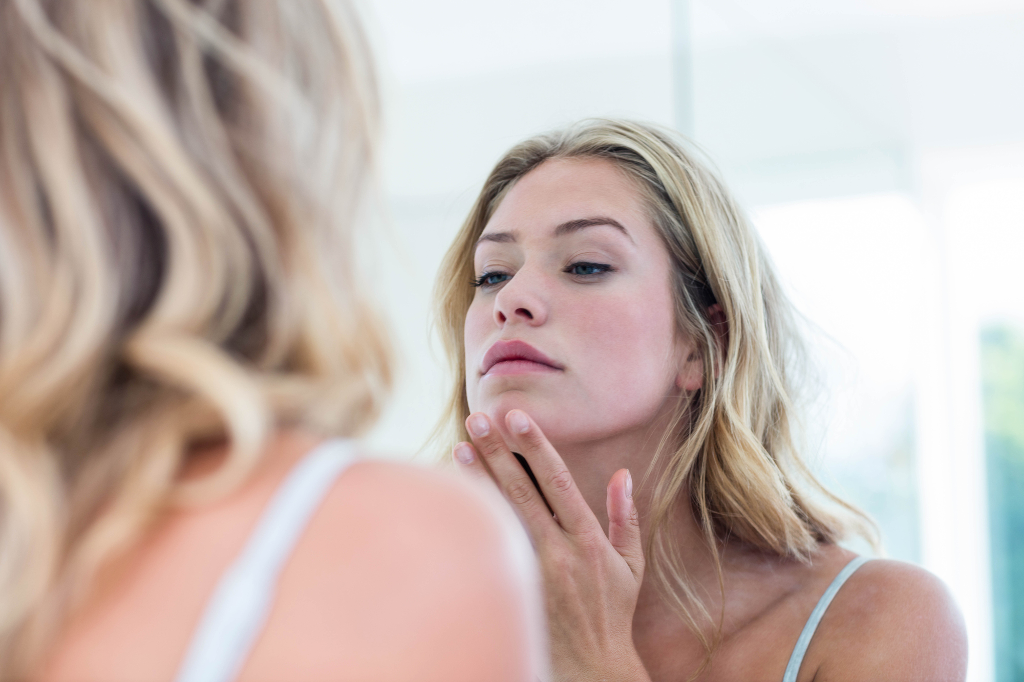 A woman with long blonde hair looks at herself in the mirror, gently touching her chin. She appears to be examining her skin, with a focused expression in a bright, softly lit room.