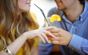 A close-up of a couple holding a glass with an orange slice garnish and a straw. The woman has long hair, wearing a yellow top, and the man has short hair, wearing a blue checkered shirt. They are smiling and looking at each other.